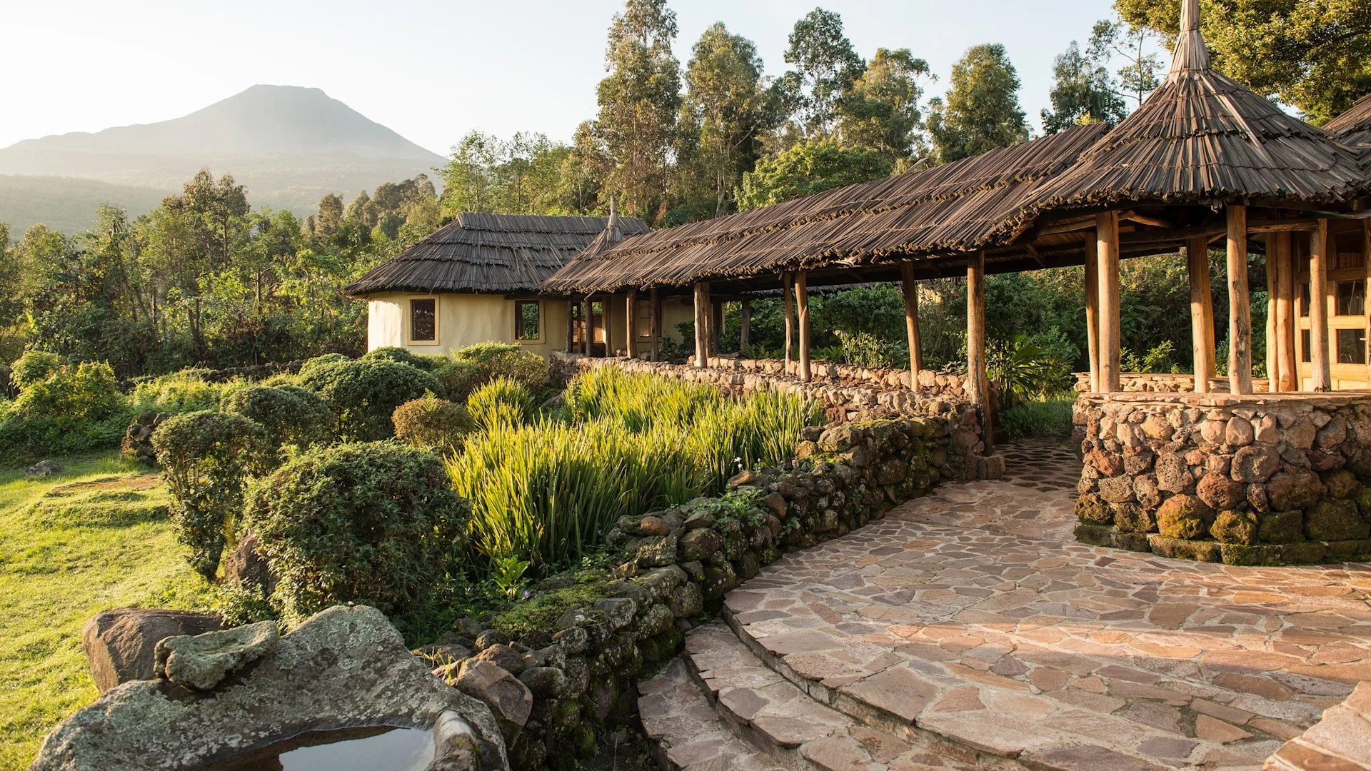 Traditional huts with thatched roofs in a lush green garden with mountains in the background.