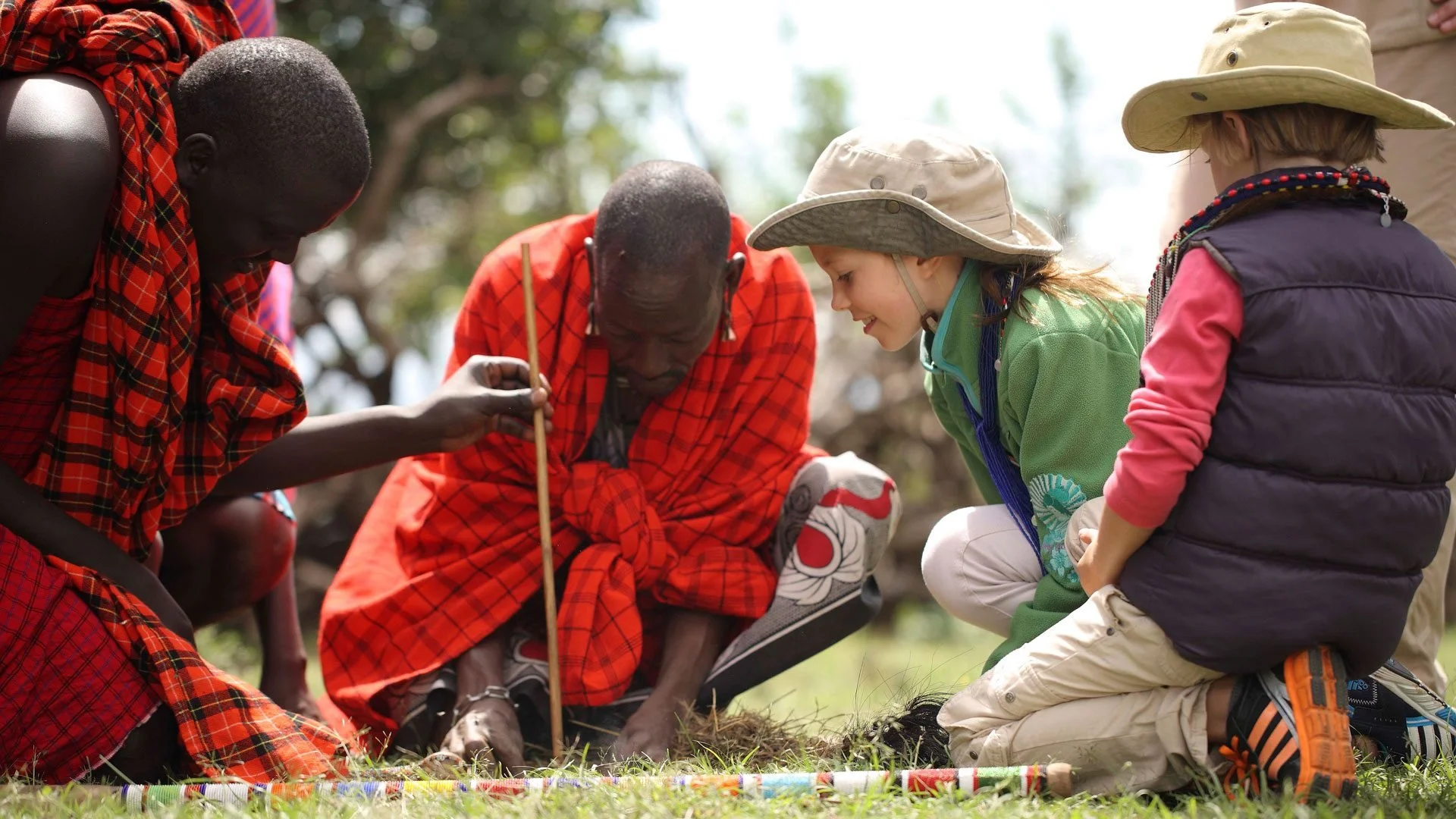 Children and an elderly man engaged in an outdoor activity, with children wearing hats and jackets, and the man dressed in traditional Maasai attire, sitting on the grass and examining something in the soil.