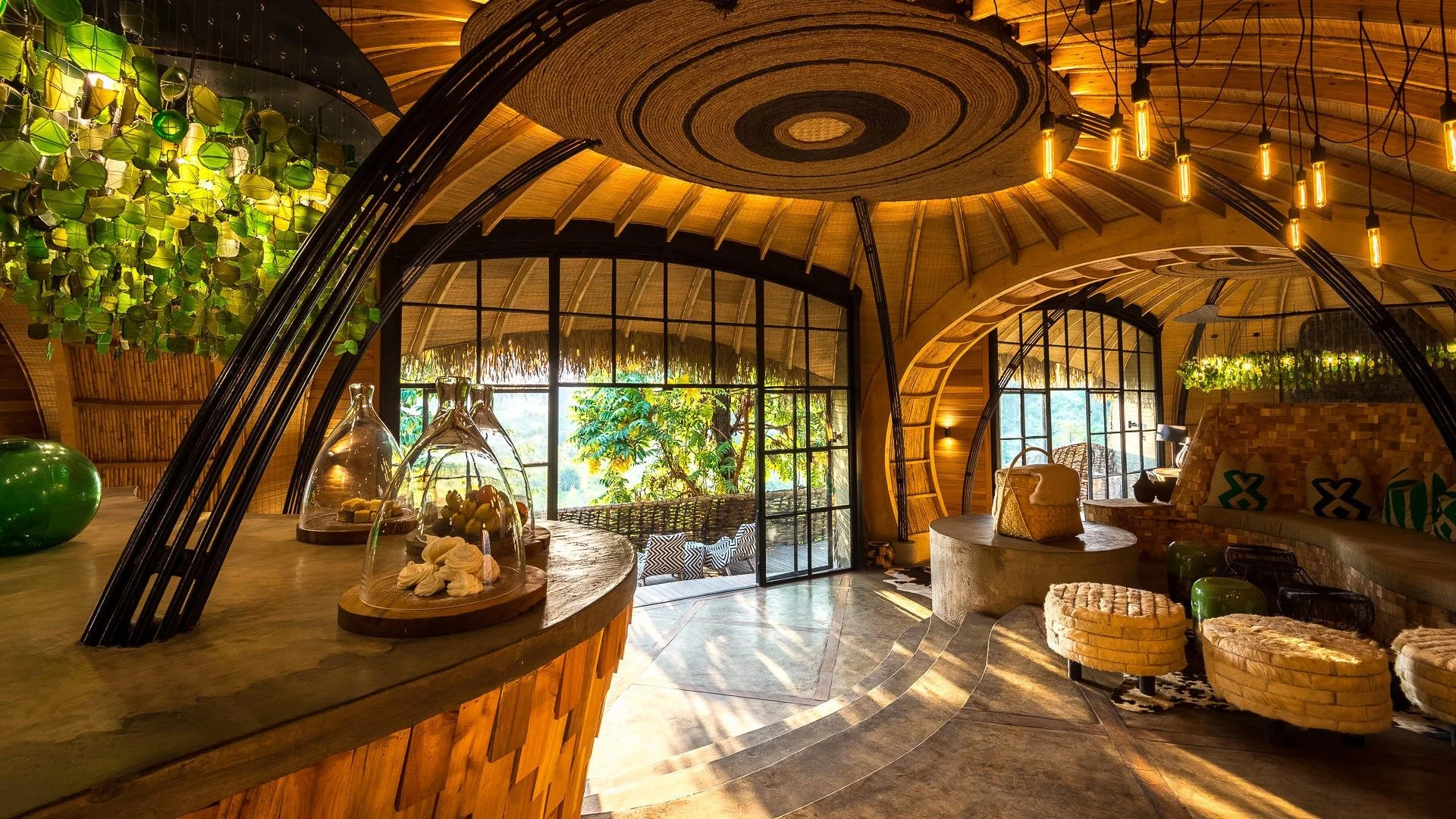 Interior of a cozy, tropical-themed lounge with curved wooden architecture, warm lighting, green plants, glass display domes with snacks, and large windows showing lush outdoor scenery.