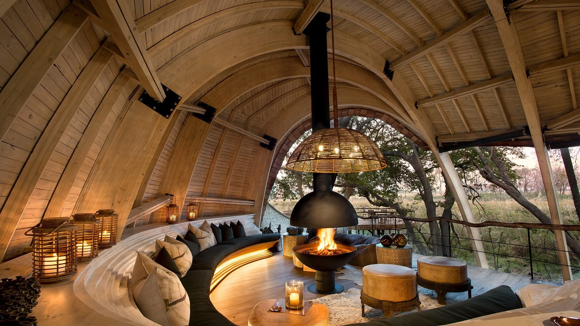 Interior of a cozy, circular treehouse with a wood ceiling and a central fireplace. There are cushions along the curved bench, candles, and decorative items, with large windows overlooking a wooded landscape outside.
