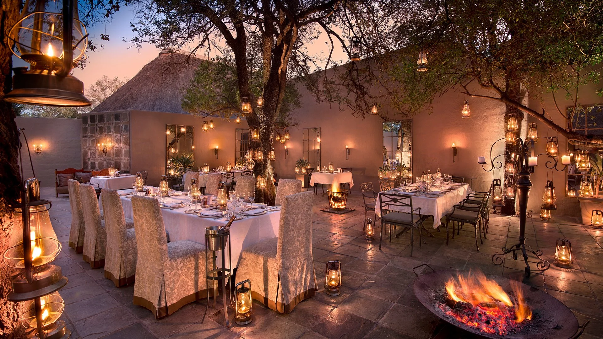 Outdoor dining area with long tables covered in white tablecloths, surrounded by upholstered chairs, lit by lanterns and wall sconces, with trees, a fire pit, and hanging lanterns at dusk.
