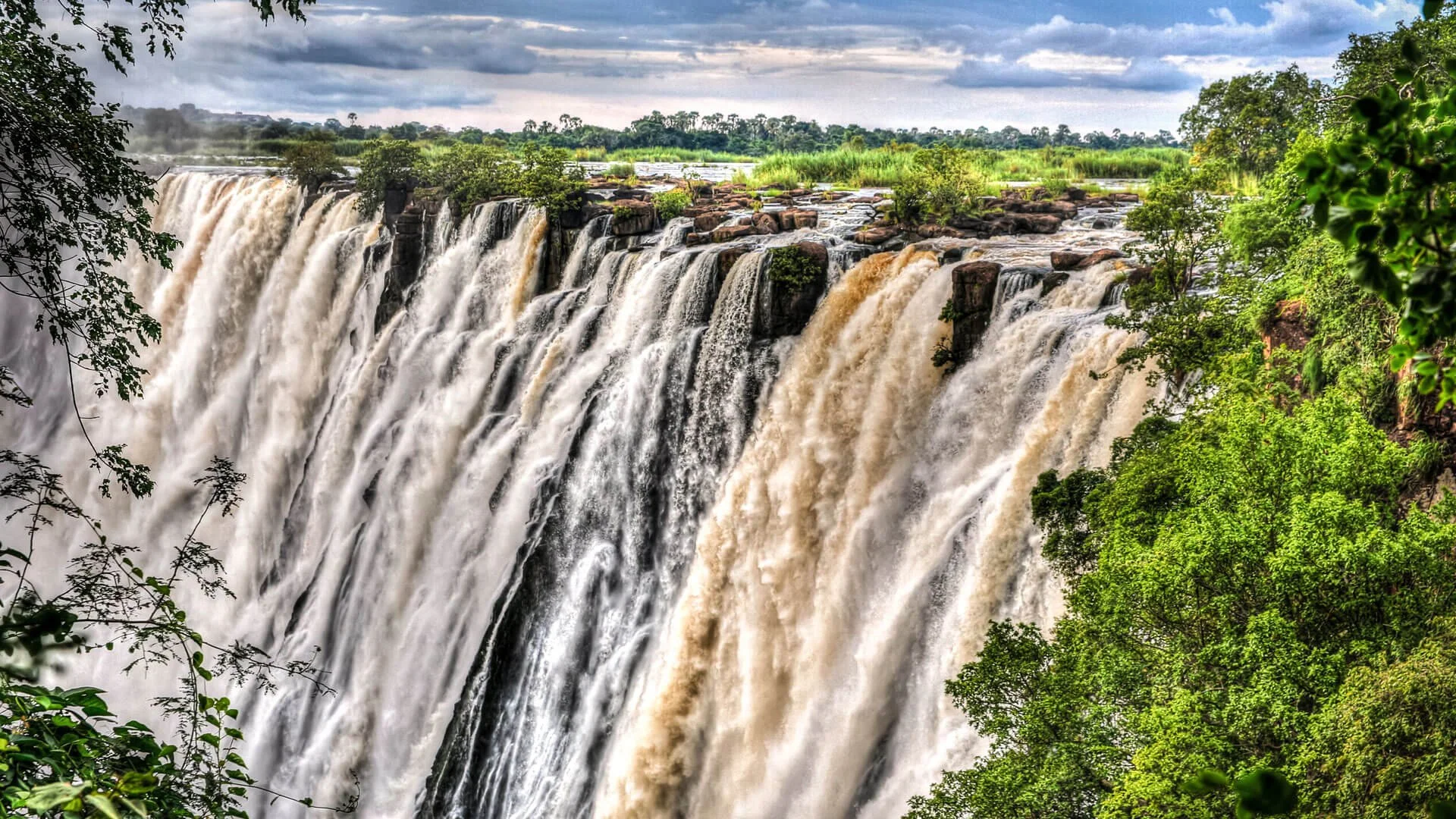 A wide view of a large waterfall cascading over rocks, surrounded by lush green trees and vegetation, with a cloudy sky overhead.