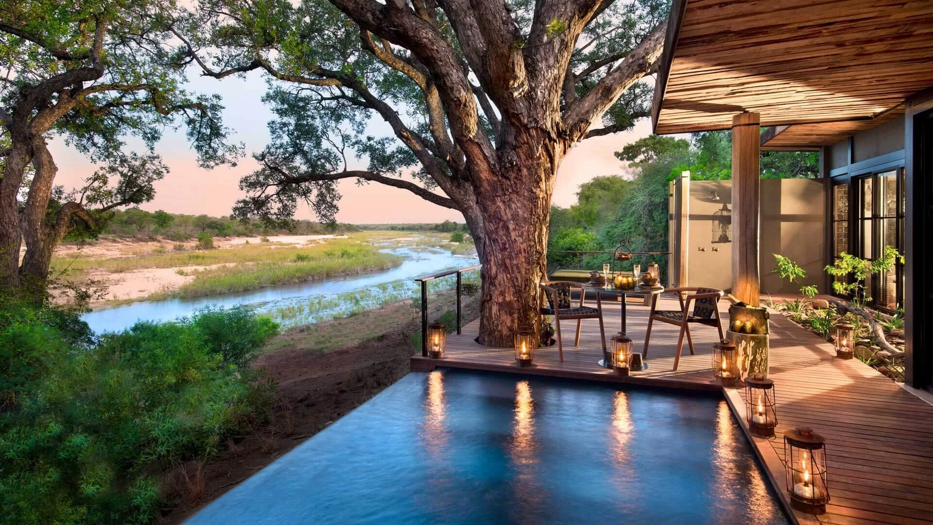 Luxury outdoor deck overlooking a river with a large tree, glass lanterns, and a dining setup under a canopy in a natural setting.