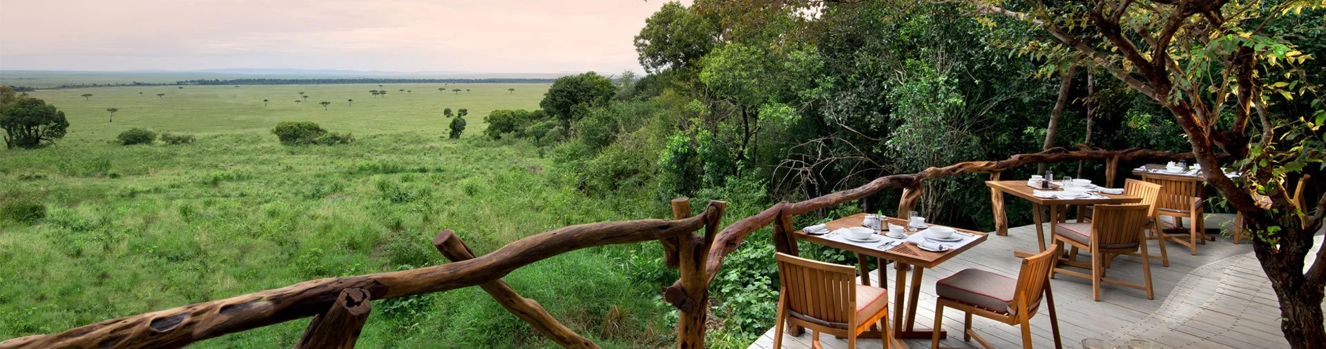 Outdoor dining area with wooden tables and chairs on a deck overlooking a grassy savannah with scattered trees and bush, under a cloudy sky.