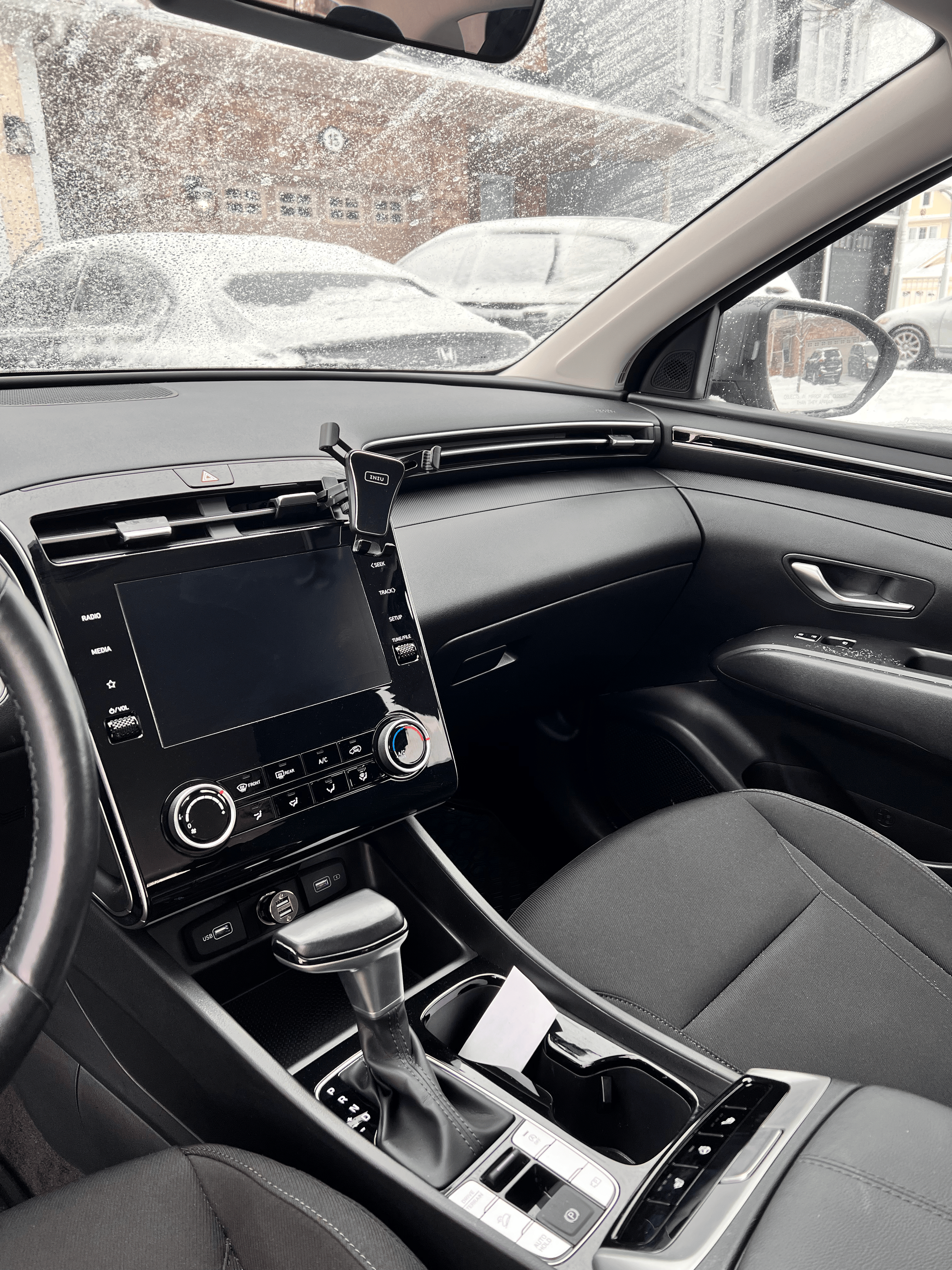 Interior of a car showing the dashboard, steering wheel, gear shift, and phone holder, with snowy cars visible outside the window.