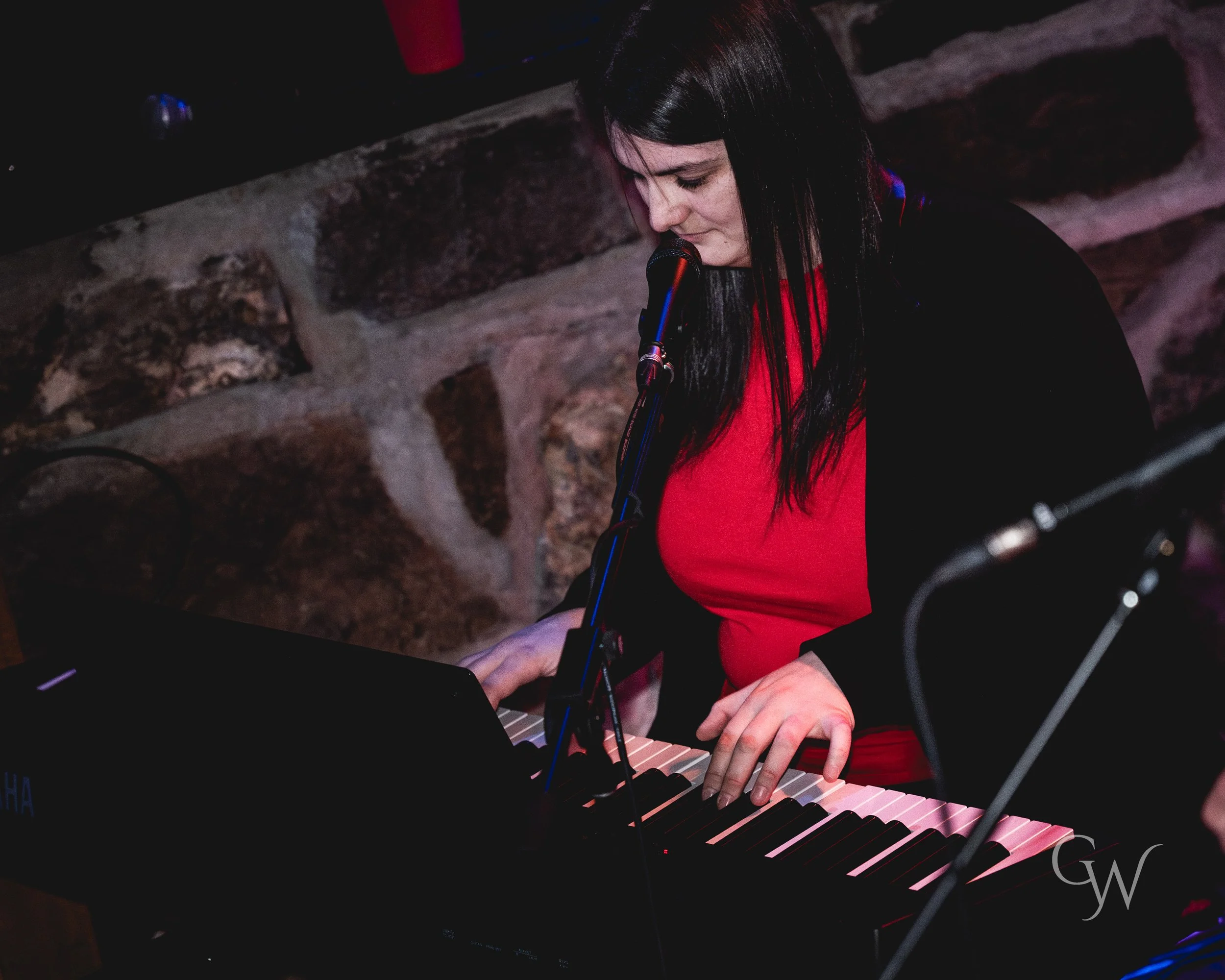 Woman with black hair and red shirt playing a piano and singing into a microphone, stone wall background.