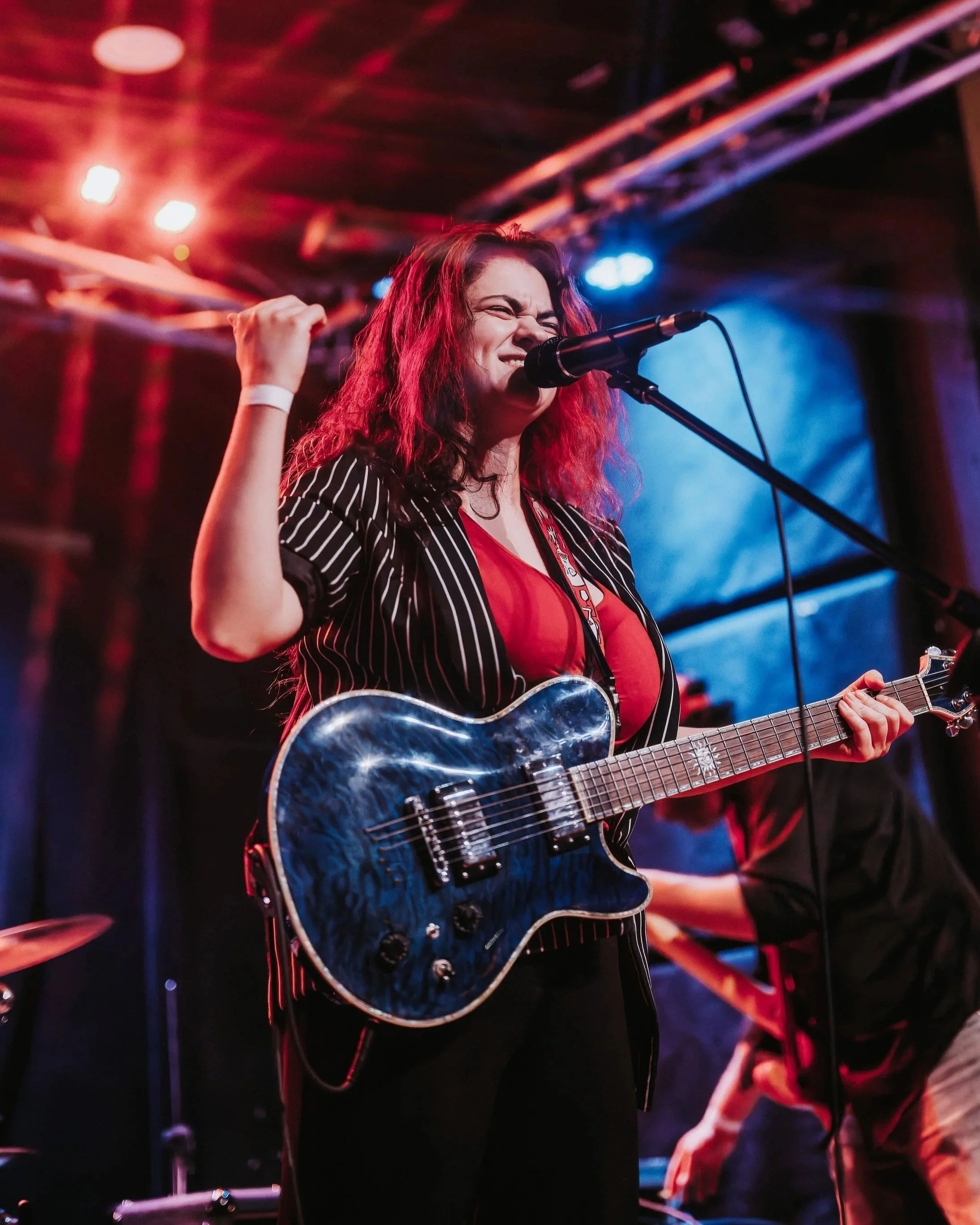 A woman with long, curly red hair singing into a microphone and playing an electric guitar on stage, with colorful stage lighting.