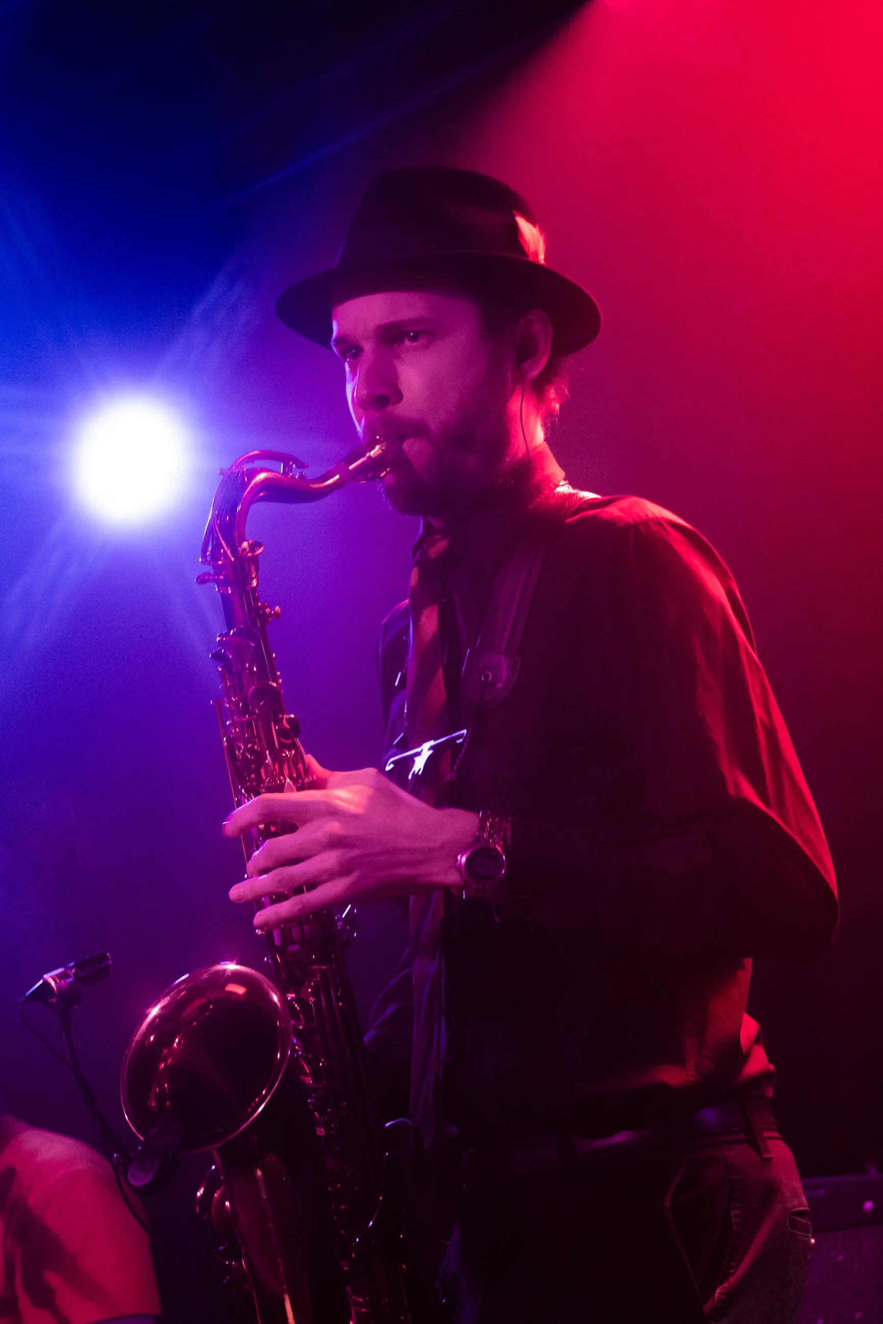 A man wearing a black hat and dark clothing playing a saxophone on stage, illuminated by purple and red stage lights.