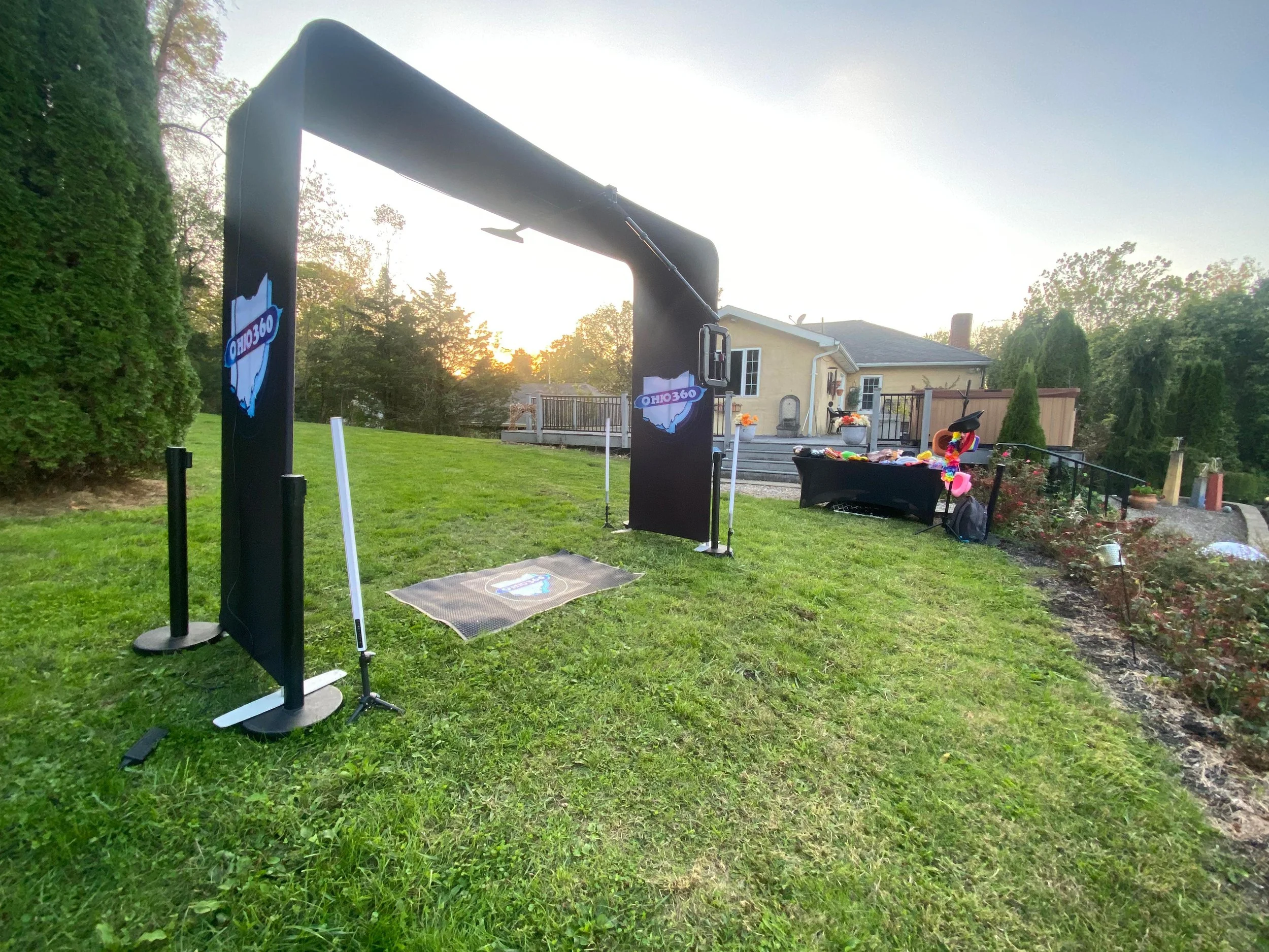 Backyard with a black inflatable archway and a photo booth area with decorations, set up for a celebration at sunset.