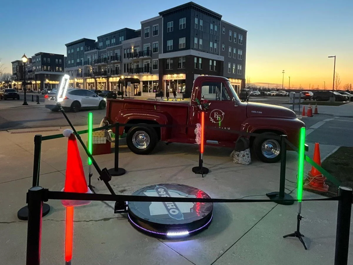 A vintage red truck is decorated for Christmas and displayed outdoors at sunset, surrounded by orange and green neon lights, orange cones, and a small barrier. A circular sign on the ground features the number 360.