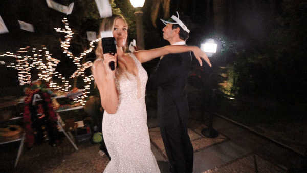 A bride and groom are dancing at their wedding reception outdoors at night. The bride is holding a microphone and wearing a white gown, while the groom is dressed in a dark suit and a paper hat. String lights are visible in the background, creating a festive atmosphere.