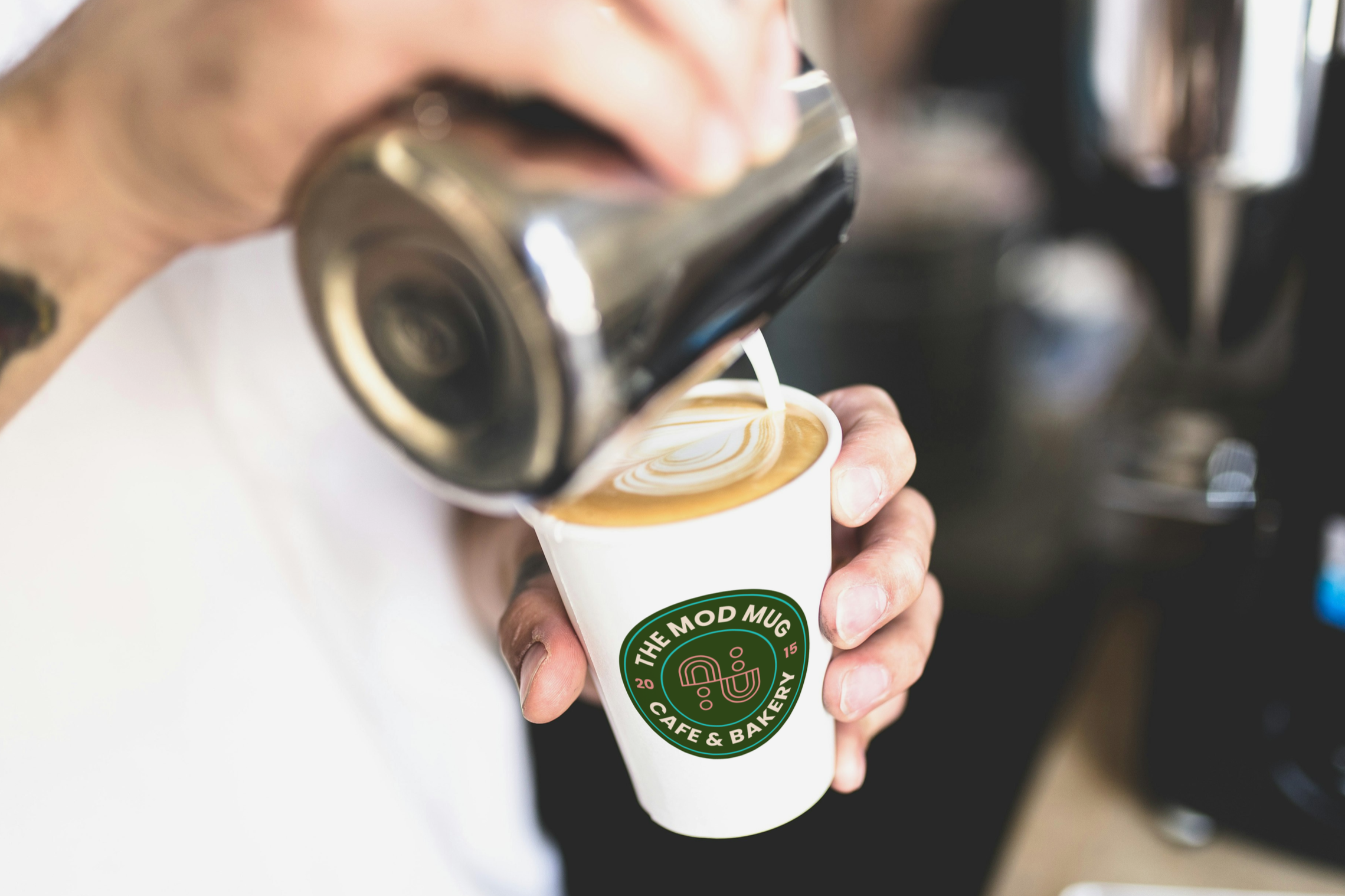 Barista pouring steamed milk into a cup of coffee at a cafe, logo of The Mod Mug Café & Bakery on the cup.
