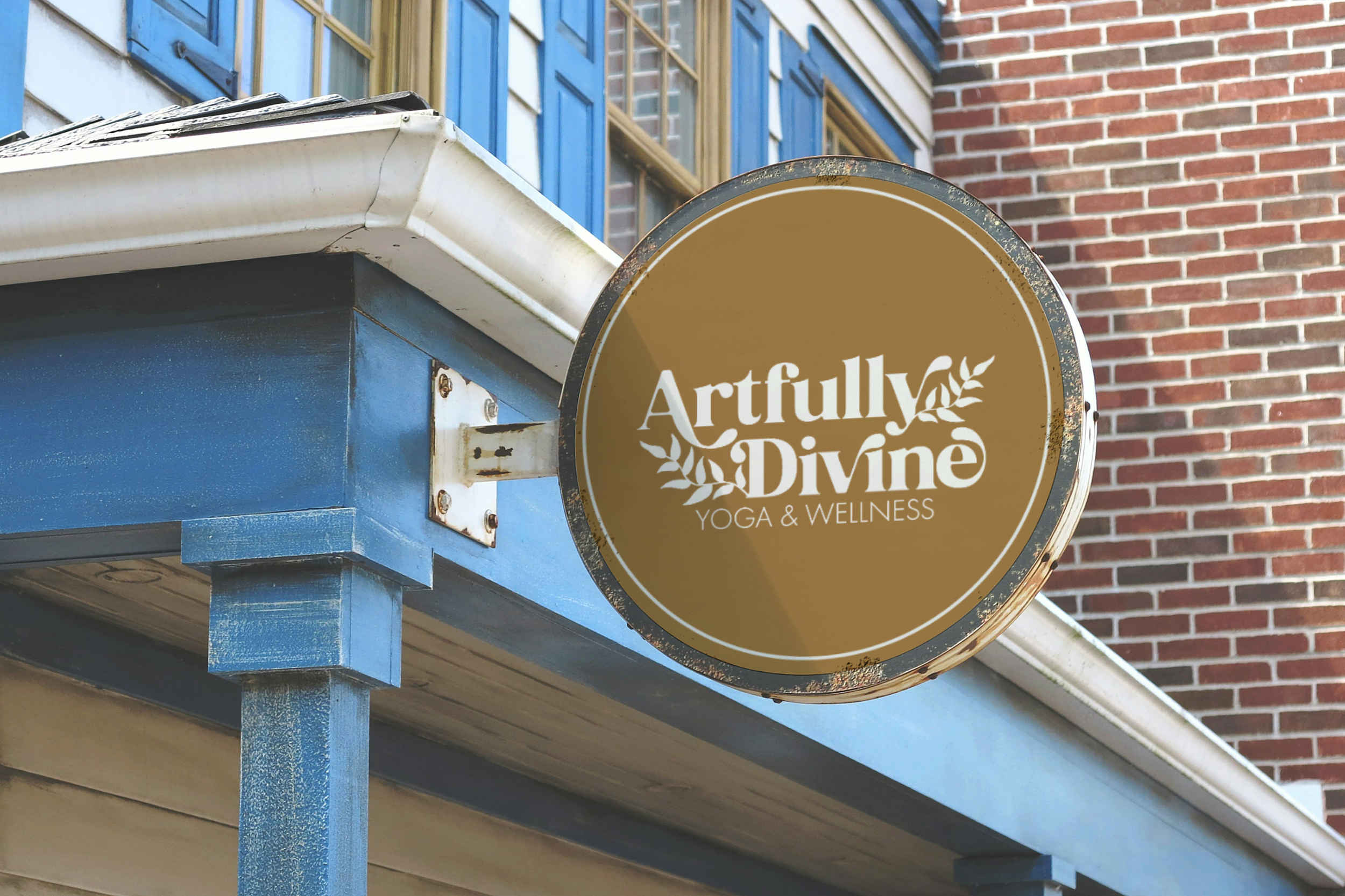 A round sign for 'Artfully Divine Yoga & Wellness' mounted on the corner of a blue building, with a background of brick wall and windows.