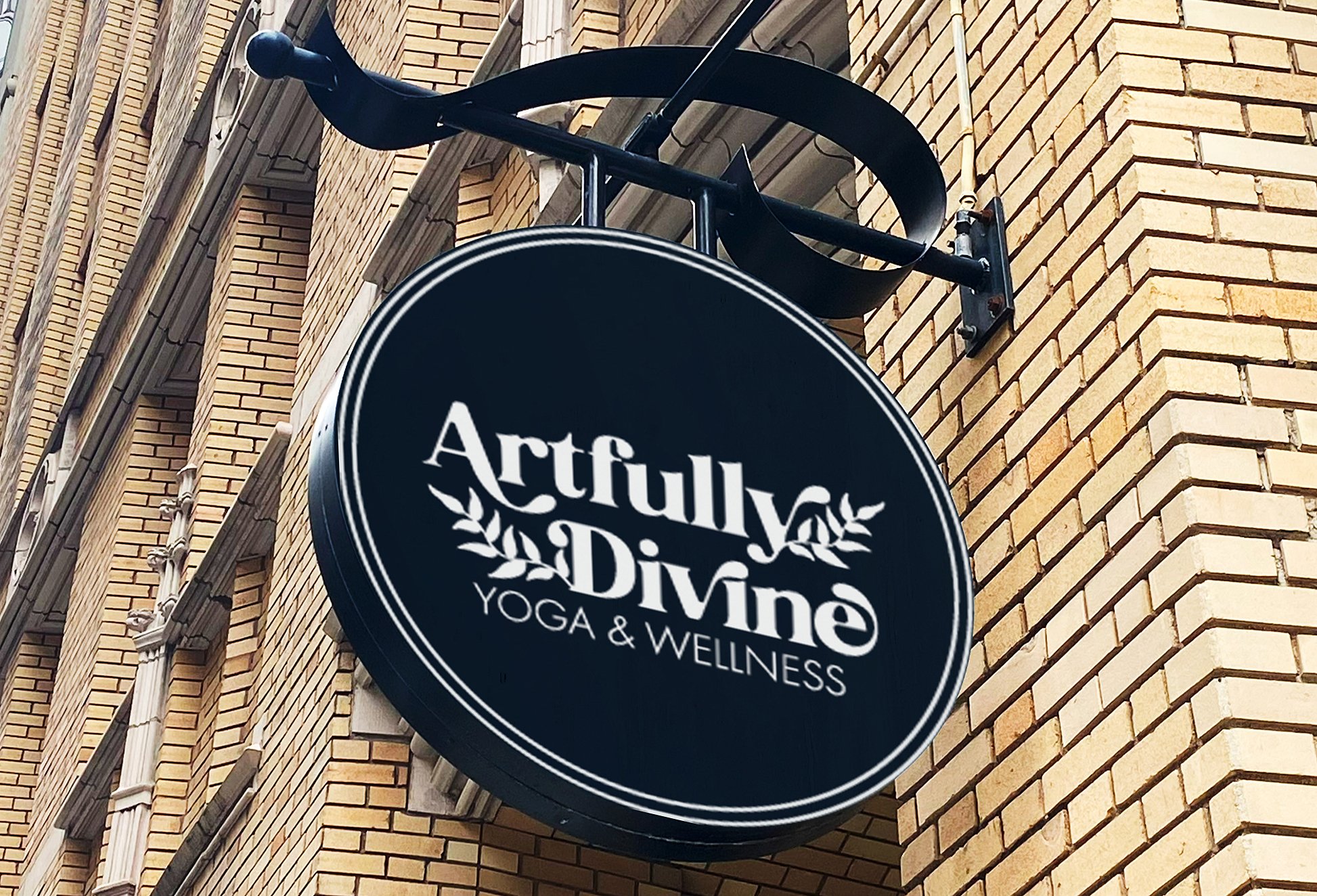Black circular sign for Artullya Divine Yoga & Wellness hanging on a brick building exterior.