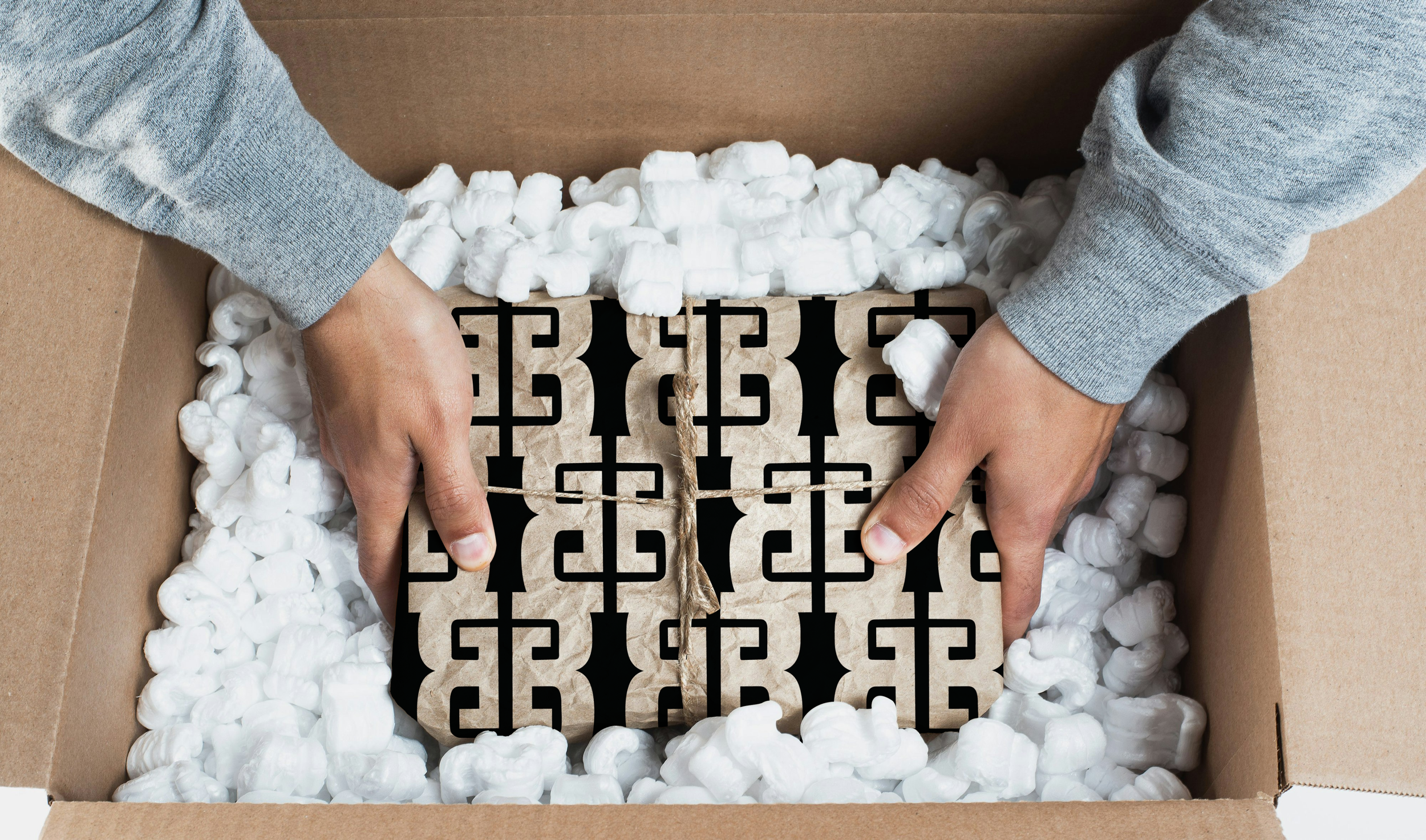 Person unboxing a decorative metal grate wrapped in brown paper, inside a cardboard box filled with packing peanuts.