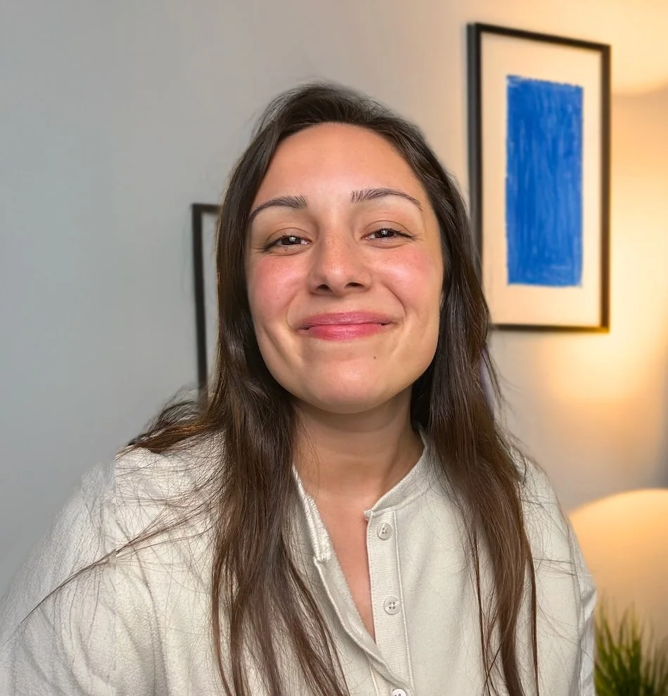 Abby Castillo with long brown hair wearing a cream-colored shirt, standing in front of a light-colored wall with framed artwork and a lamp.