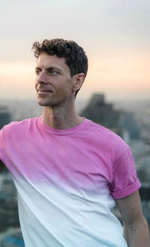 Mann in pink-weißem T-Shirt mit städtischem Hintergrund bei Sonnenuntergang.