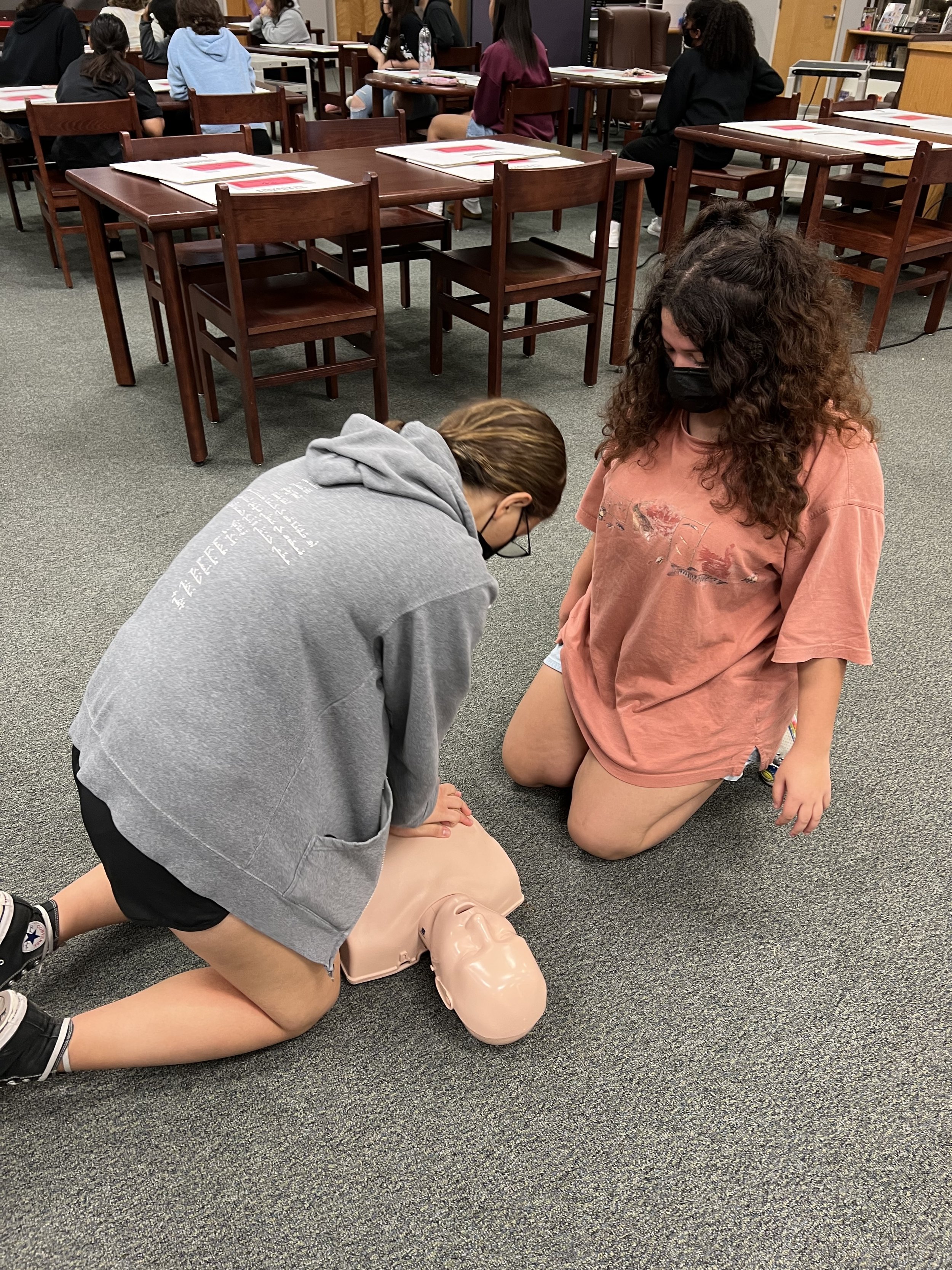 Two girls practicing CPR on a training mannequin in a classroom setting with tables and chairs.