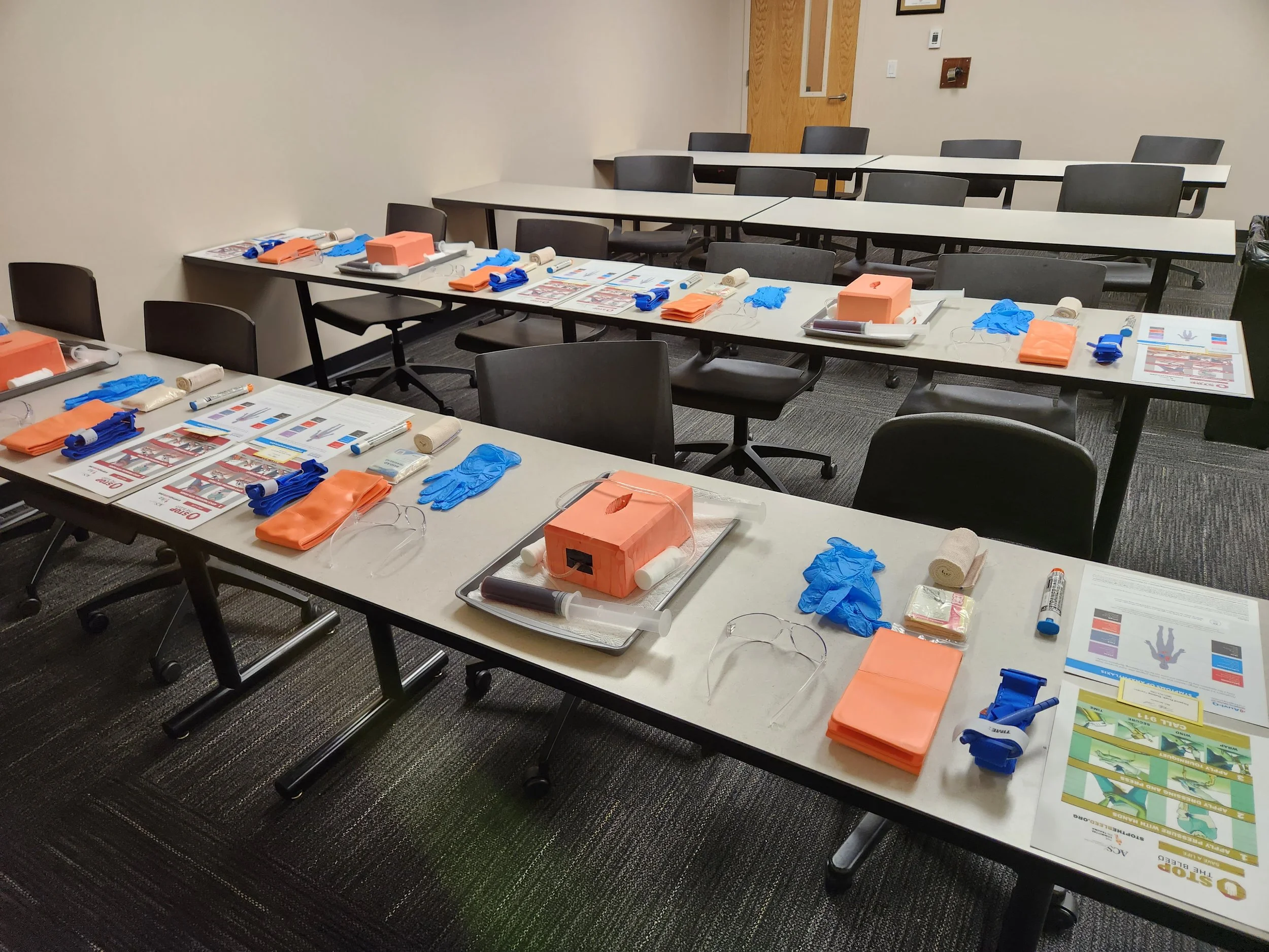 Classroom with medical training supplies laid out on tables, including gloves, bandages, face shields, and instruction sheets, ready for training session.