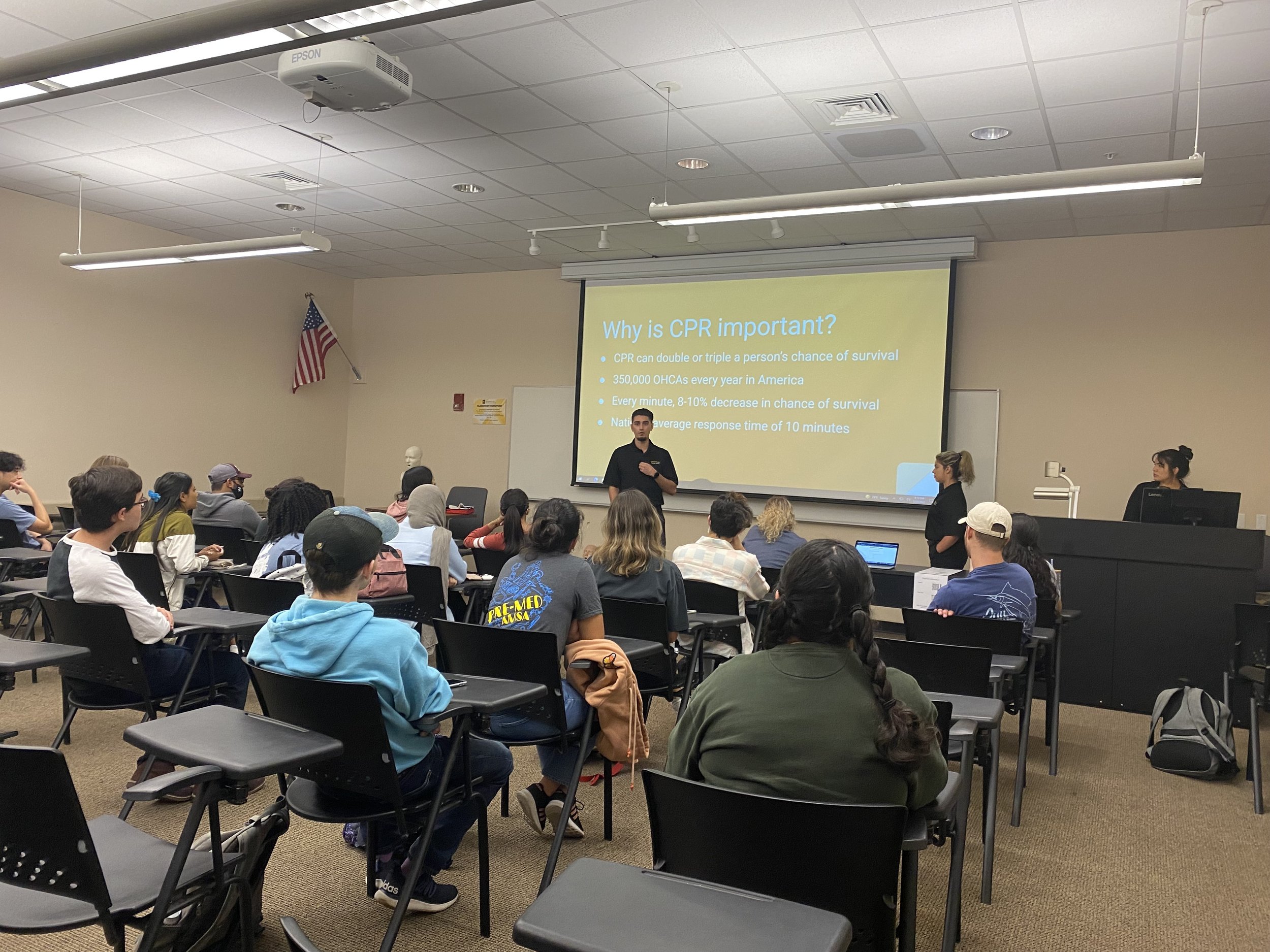 A classroom with students attending a CPR training session. A male instructor stands in front of a large screen with a presentation titled 'Why is CPR important?' and points to the text. The classroom has a white ceiling with air vents and a projecto
