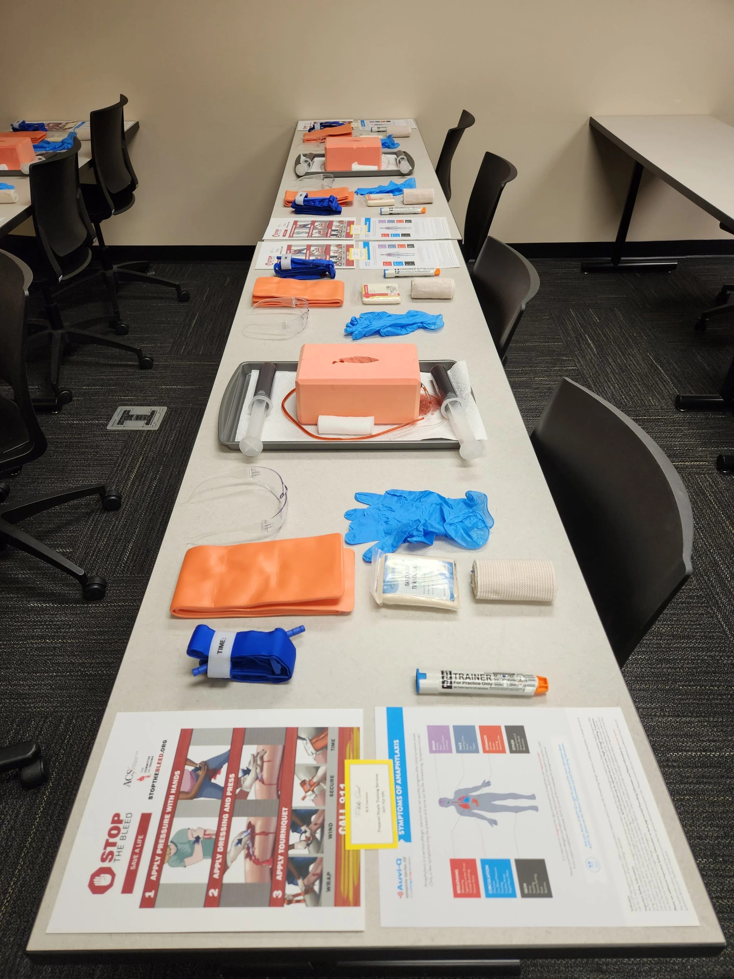 Emergency medical training supplies arranged on a long table, including gloves, defibrillator pads, syringes, instructional sheets, and a mannequin for practice.