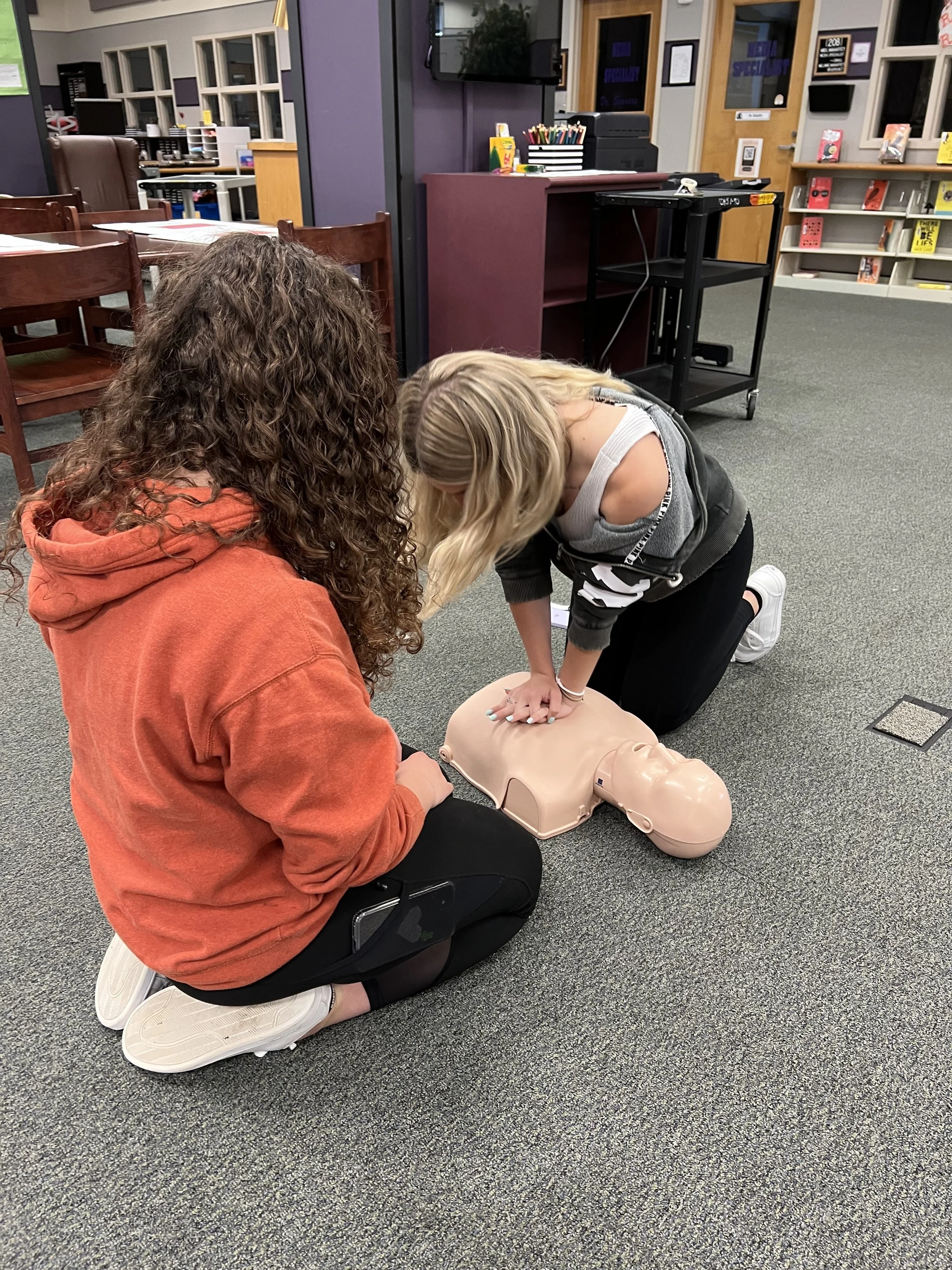 Two women practicing CPR on a mannequin in a classroom setting.