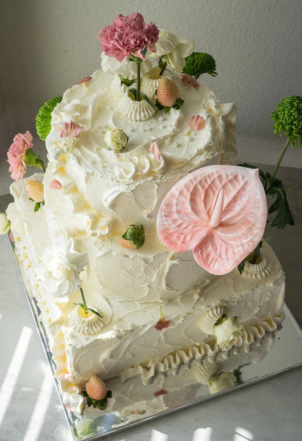 A three-tiered white wedding cake decorated with pink and white flowers, green foliage, and an anthurium flower on the front.