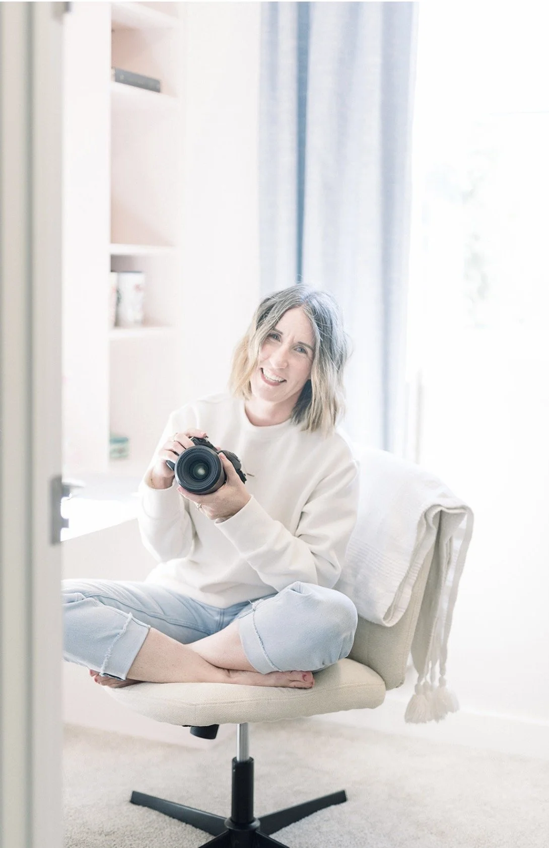 A woman with shoulder-length blonde hair sitting cross-legged on an office chair, holding a camera and smiling at the mirror. She is wearing a white sweatshirt and light blue jeans. There is a white towel draped over the back of the chair. The background features bright natural light coming through a window with blue curtains and a white shelf.