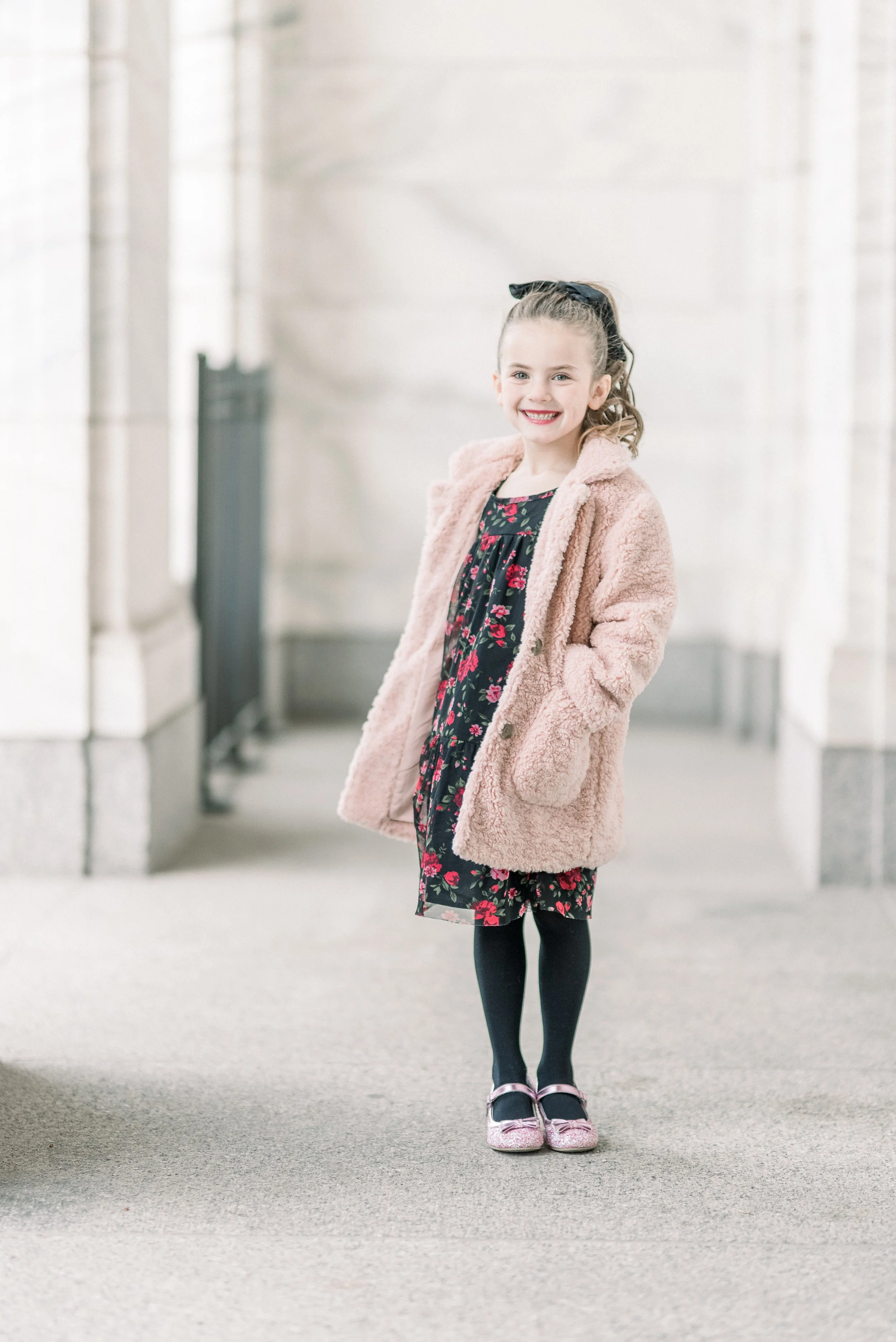 A young girl smiling, wearing a black dress with a red floral pattern, black tights, pink shoes, and a pink furry coat, standing in an indoor space with stone walls.
