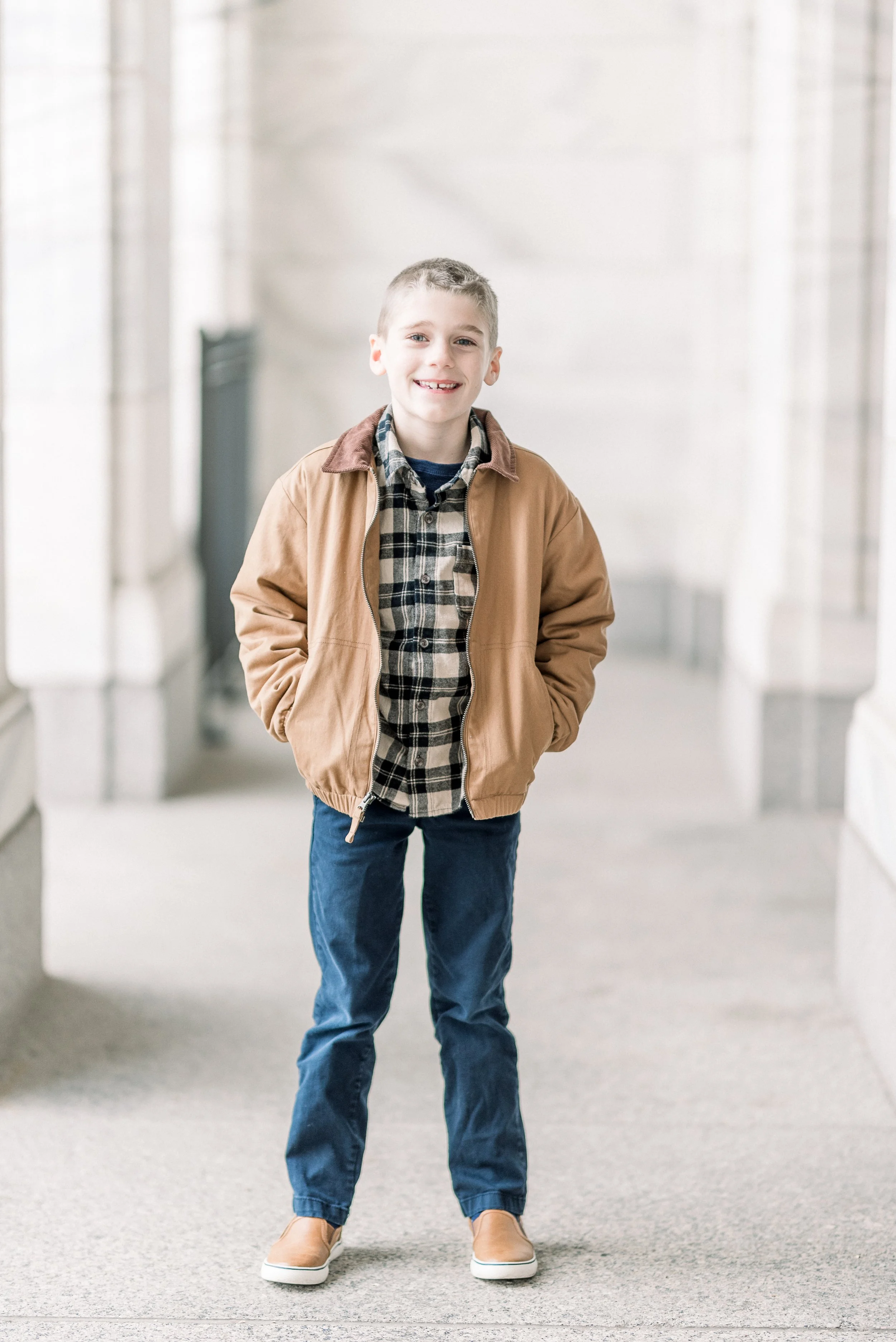 A young boy standing in a hallway with a smile, wearing a tan jacket, plaid shirt, blue jeans, and tan sneakers.