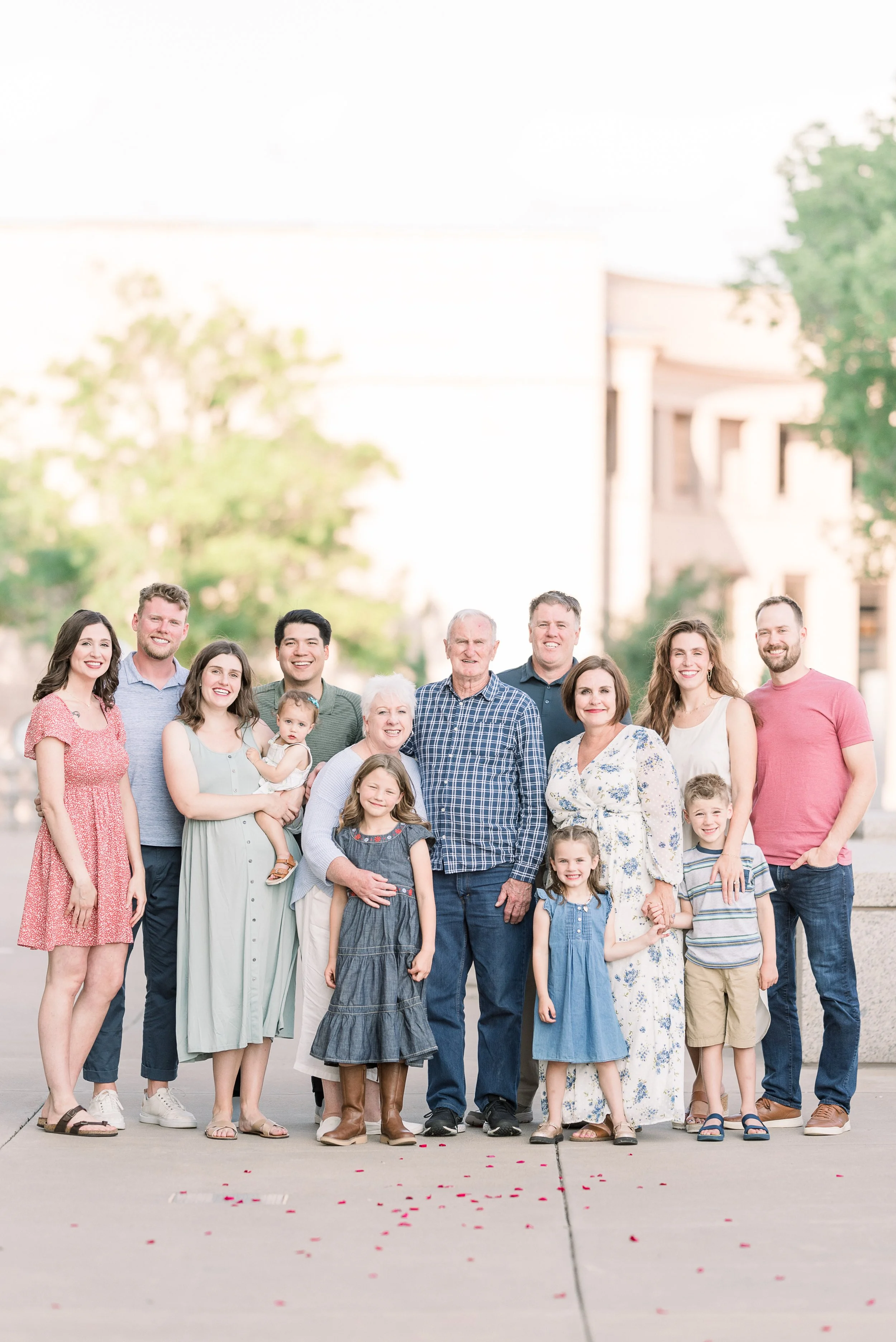 Group of fifteen people, including children and seniors, posing outdoors in front of a tree and building, smiling for a family photo.