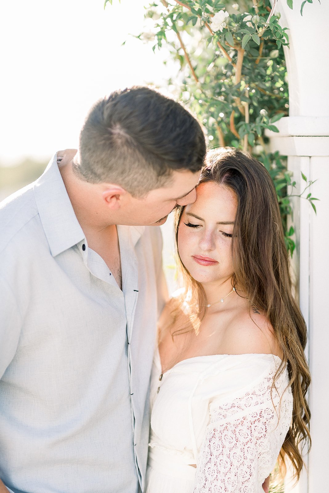 A couple sharing an intimate moment outdoors, with the man gently kissing the woman's forehead as she closes her eyes.