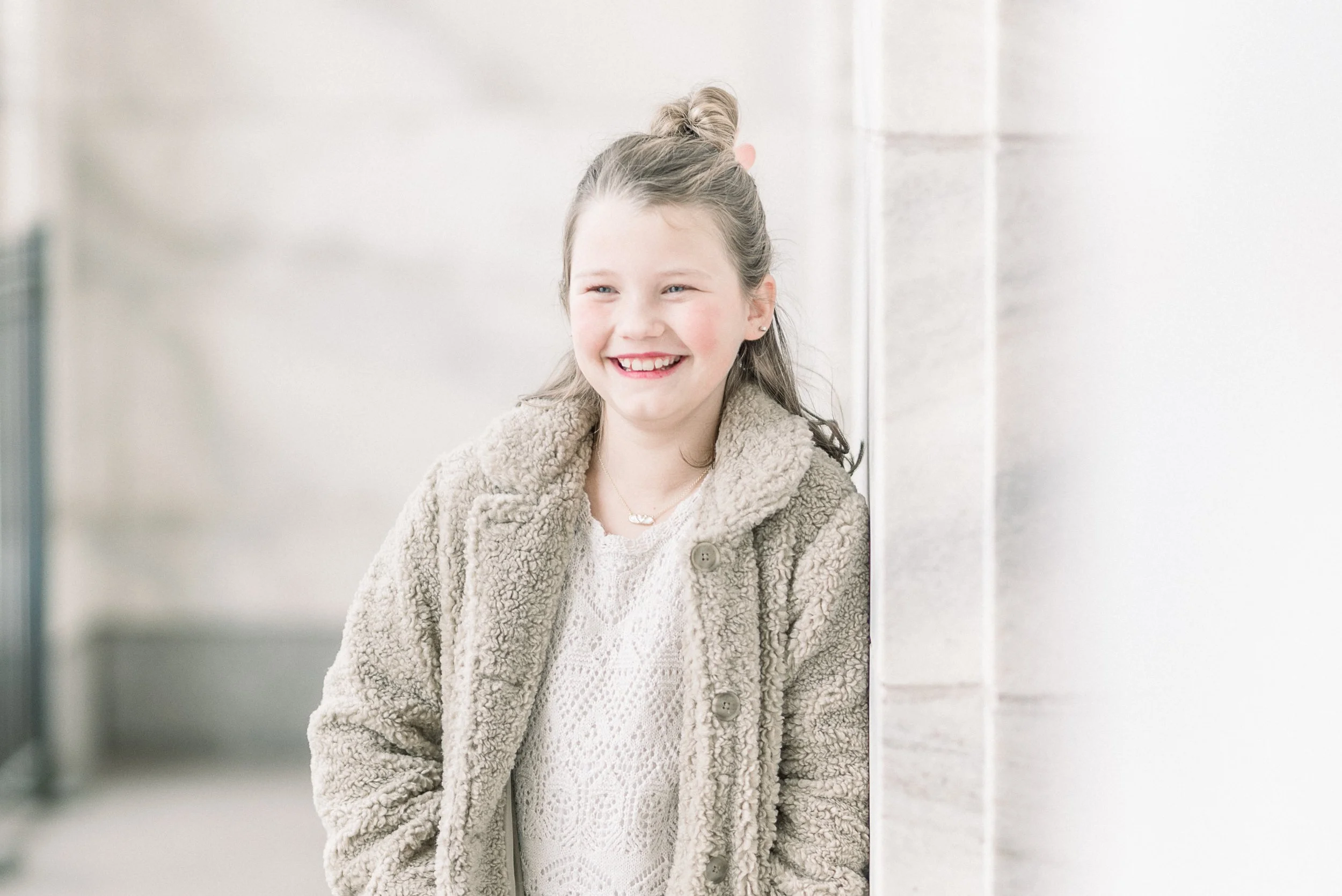 Smiling young girl with light brown hair tied up in a bun, wearing a cream-colored sweater and beige fur coat, standing against a wall in a well-lit indoor setting.