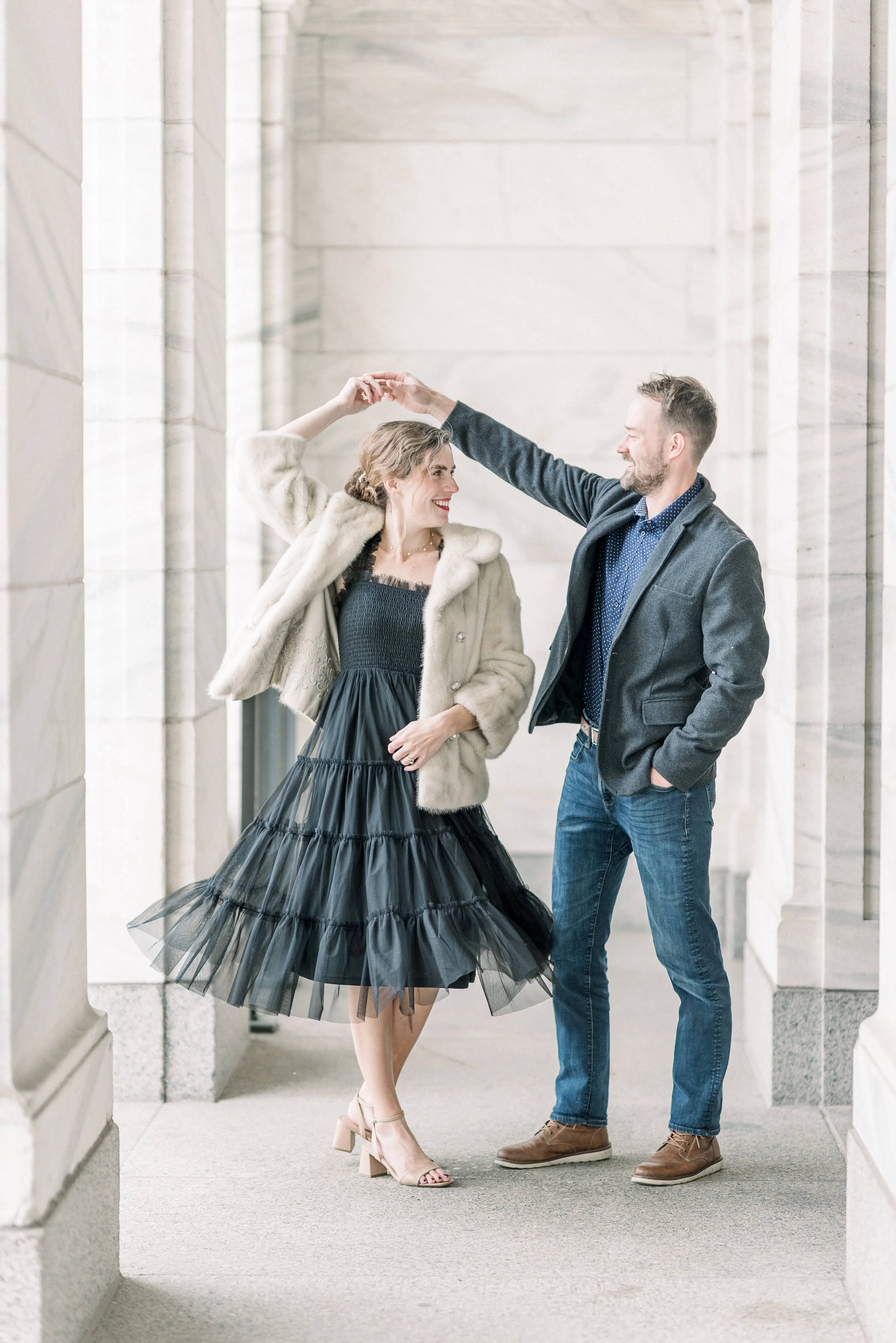 A couple dancing and smiling in a corridor with light-colored stone walls, the woman in a black dress and beige fur coat, and the man in a gray blazer and blue jeans.