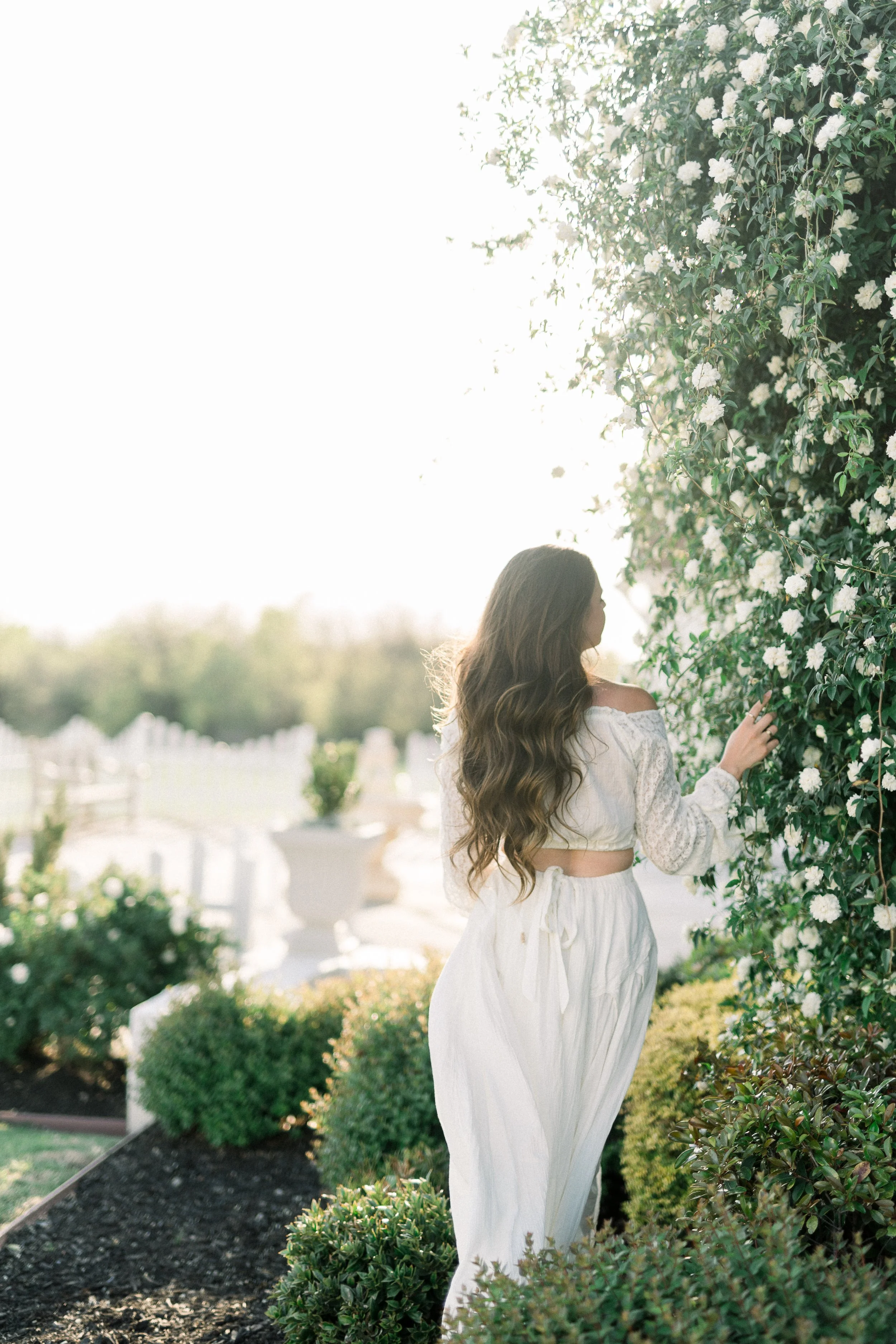 Woman with long wavy hair wearing a white off-shoulder top and white pants, touching a large white-flowered bush in a garden.