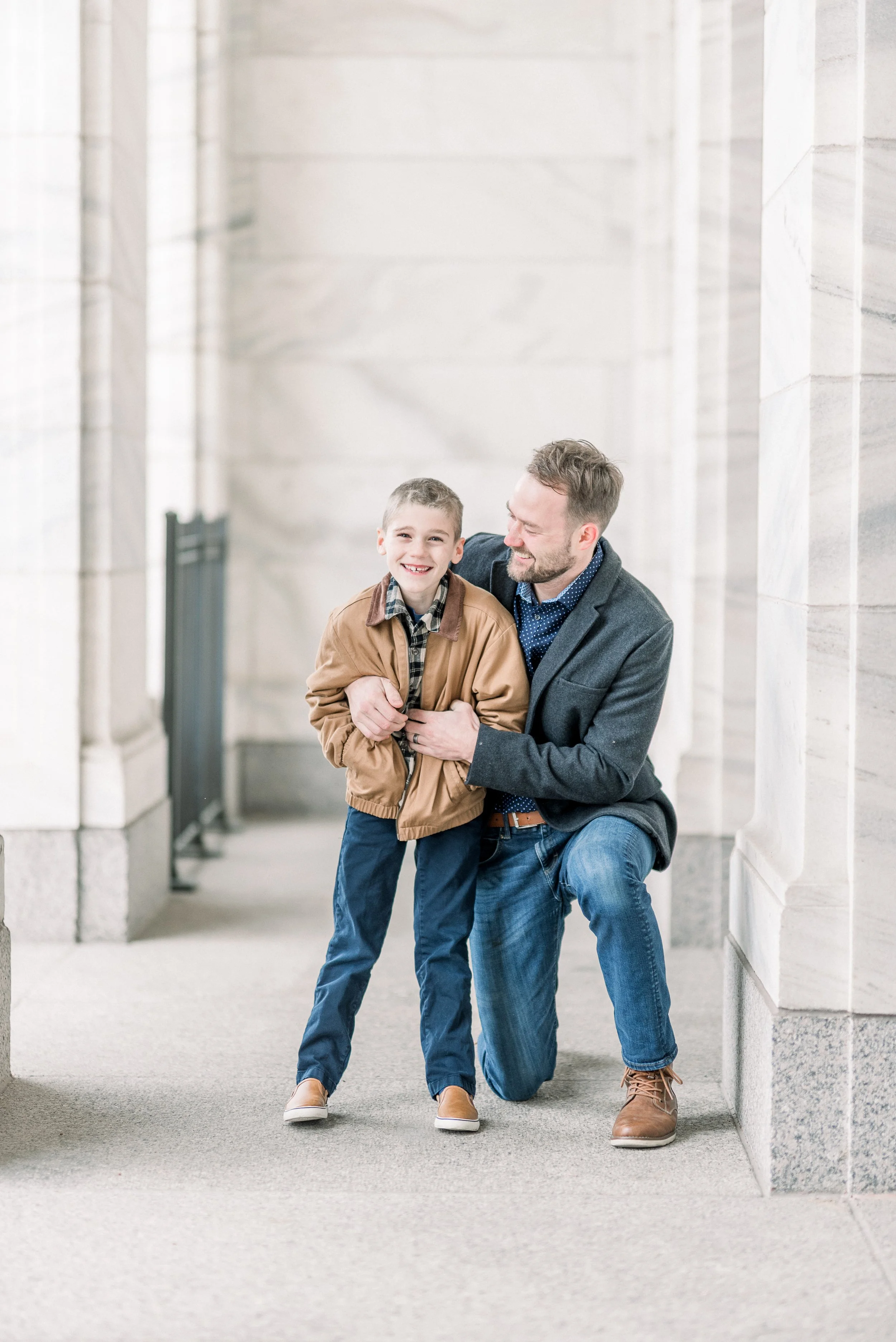 A man helping a young boy walk inside a building with marble walls, both smiling and laughing.