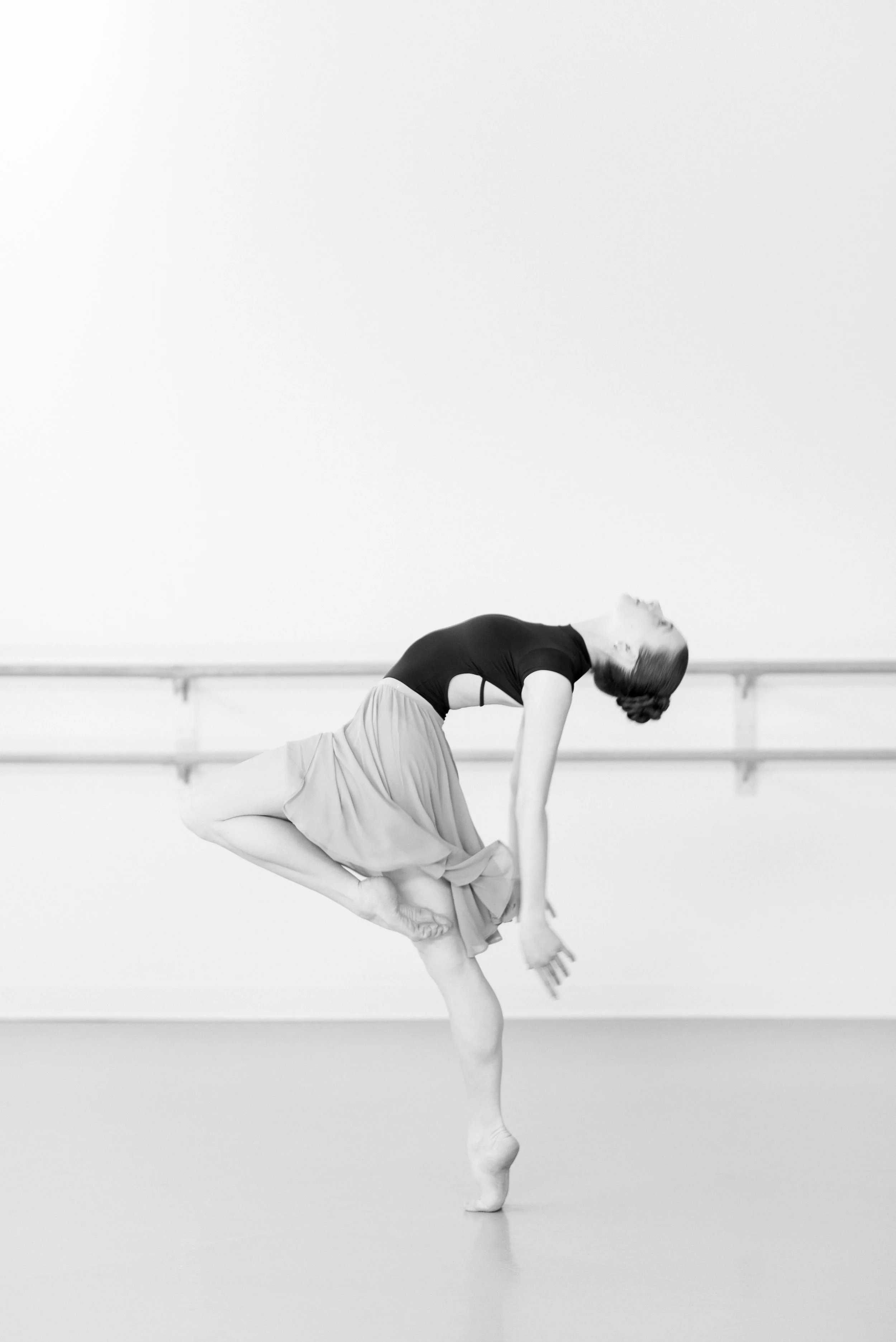 A ballet dancer in a black top and a flowing skirt performing an acrobatic pose in a dance studio.
