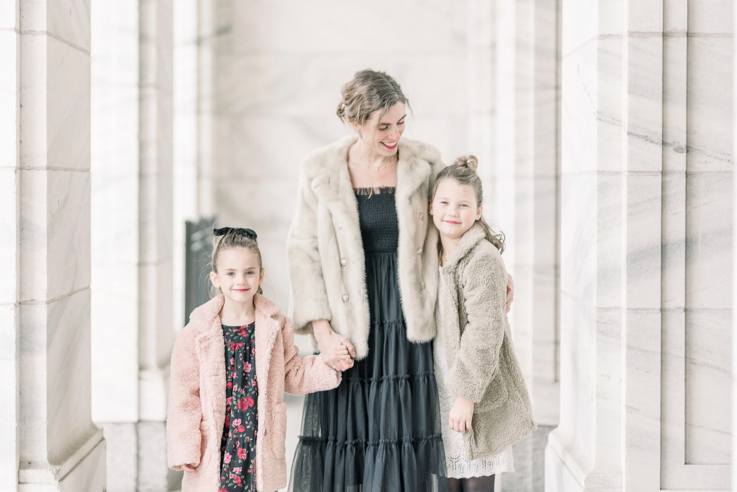 A woman with two young girls standing between stone columns. The woman is holding hands with the girl on her left and has her arm around the girl on her right, all smiling at the camera.