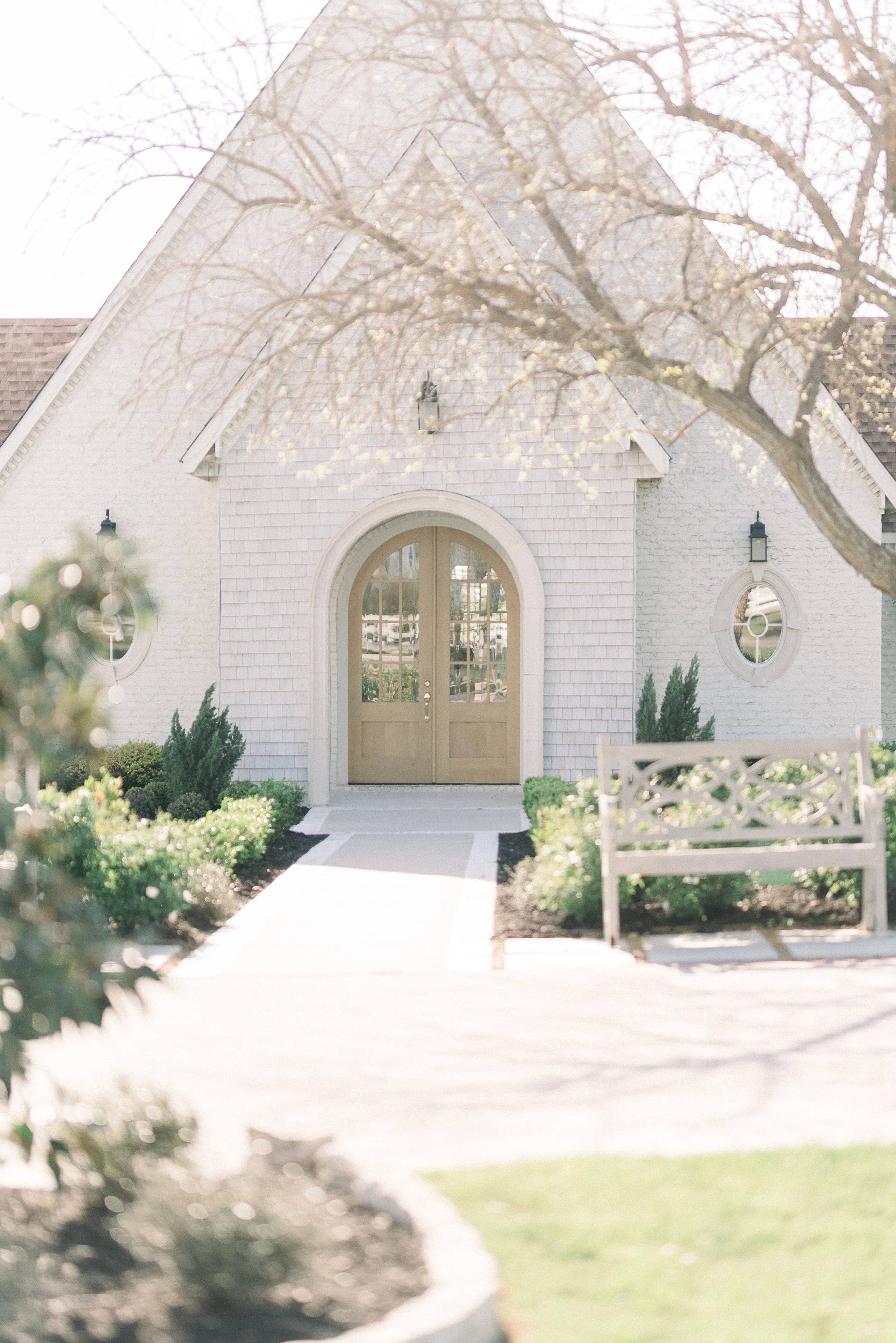A white brick building with a rounded front door and two small round windows. There is a sidewalk leading to the door, surrounded by green bushes and small trees, with a wooden bench to the right of the sidewalk. Overhead, a tree with budding leaves partially obscures the view.