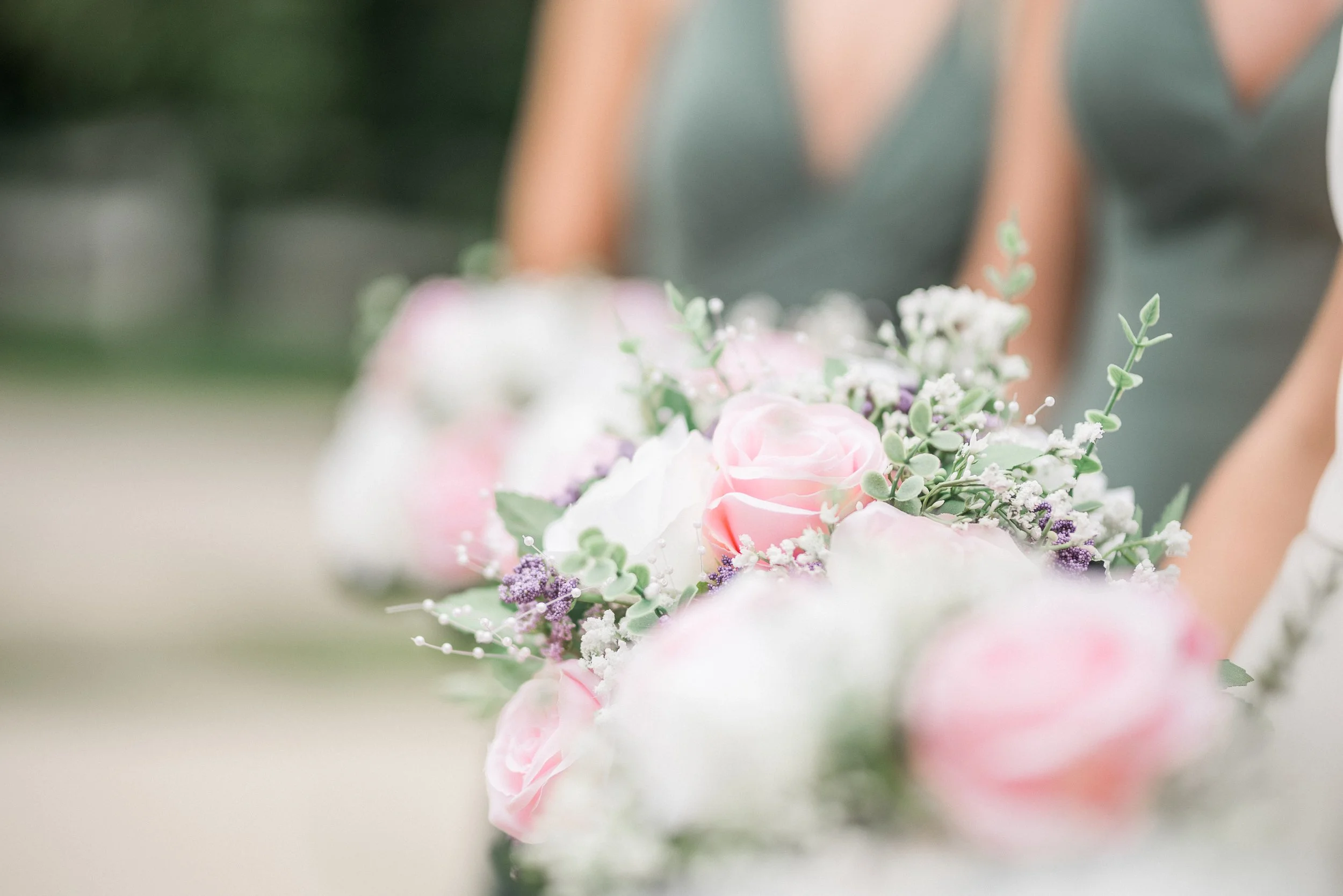A bouquet of pink and white roses with purple and green accents, held by someone in a formal outfit.