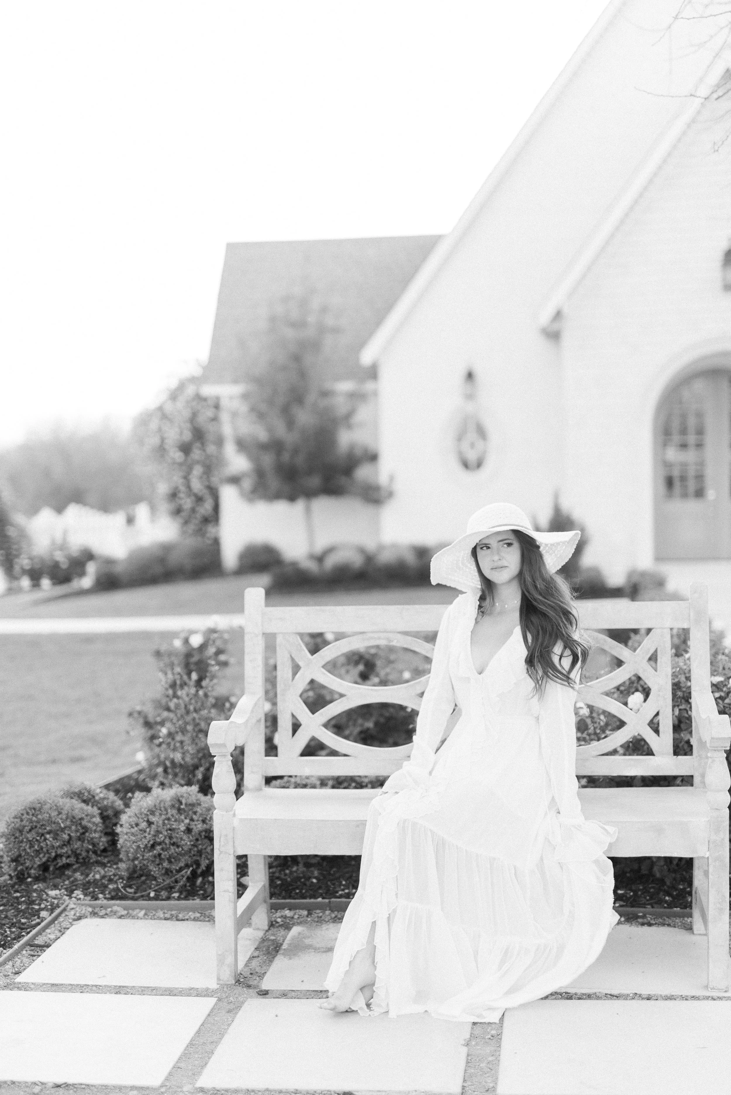 A young woman in a long white dress and wide-brimmed hat sitting on a garden bench outside a house.