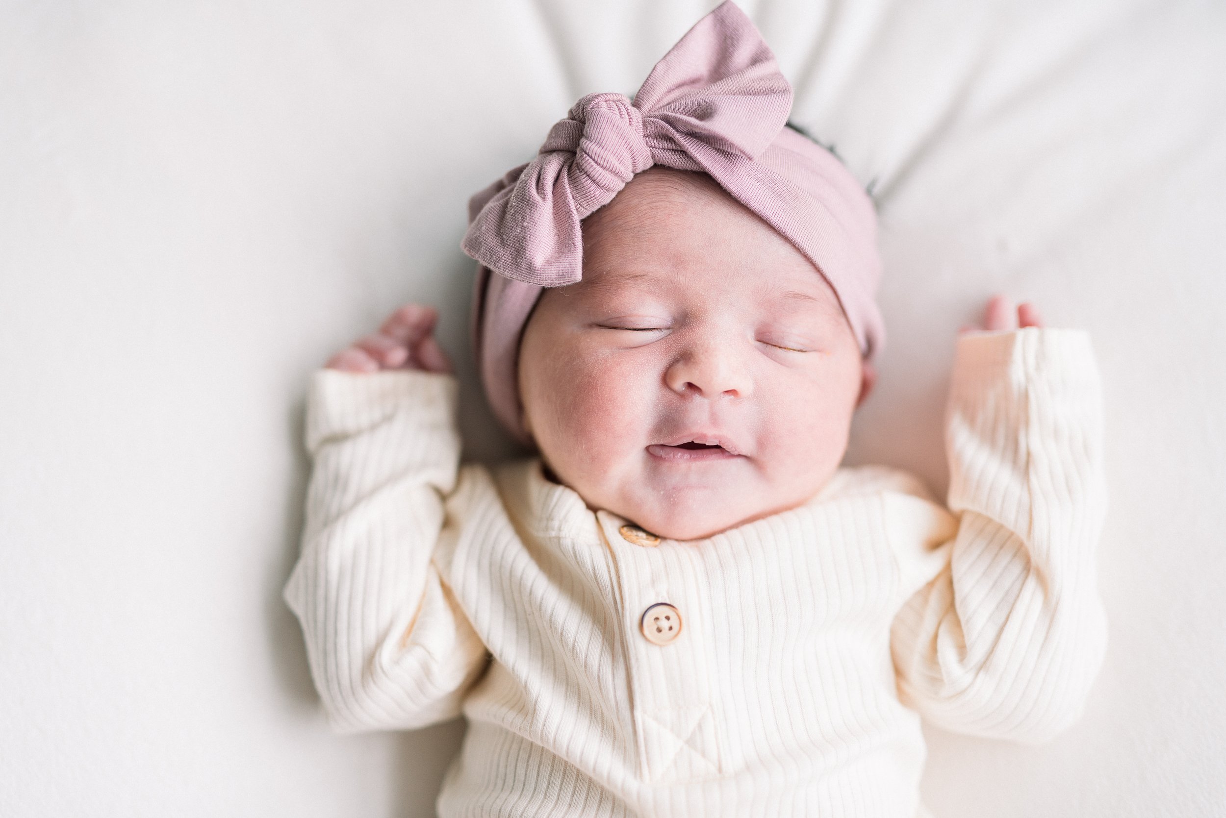 A smiling baby girl with closed eyes, wearing a pink headwrap with a bow and a cream-colored romper with buttons, lying on a white surface.
