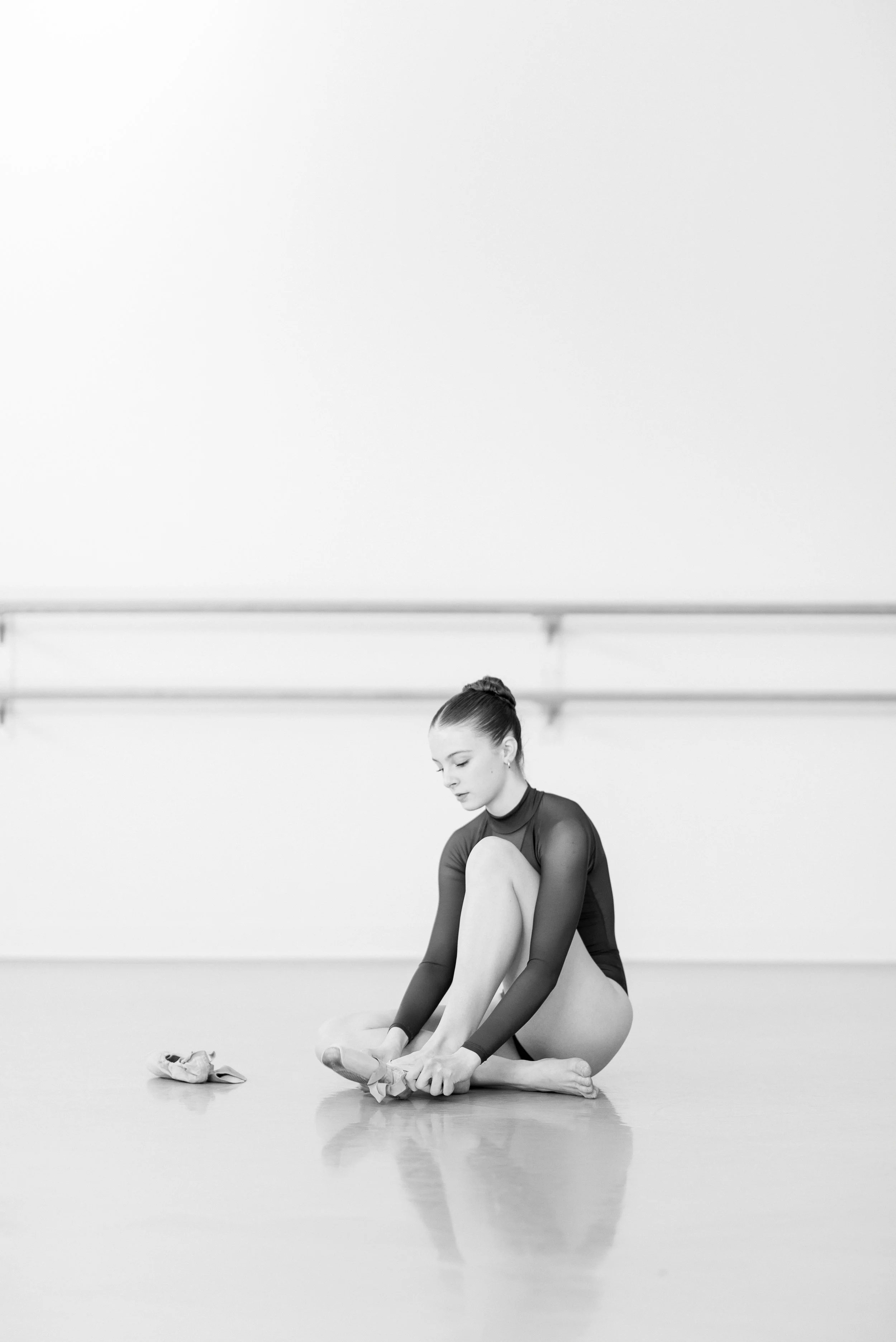 A young female ballet dancer in a dance studio, sitting on the floor, adjusting her ballet slippers, with a ballet barre in the background.