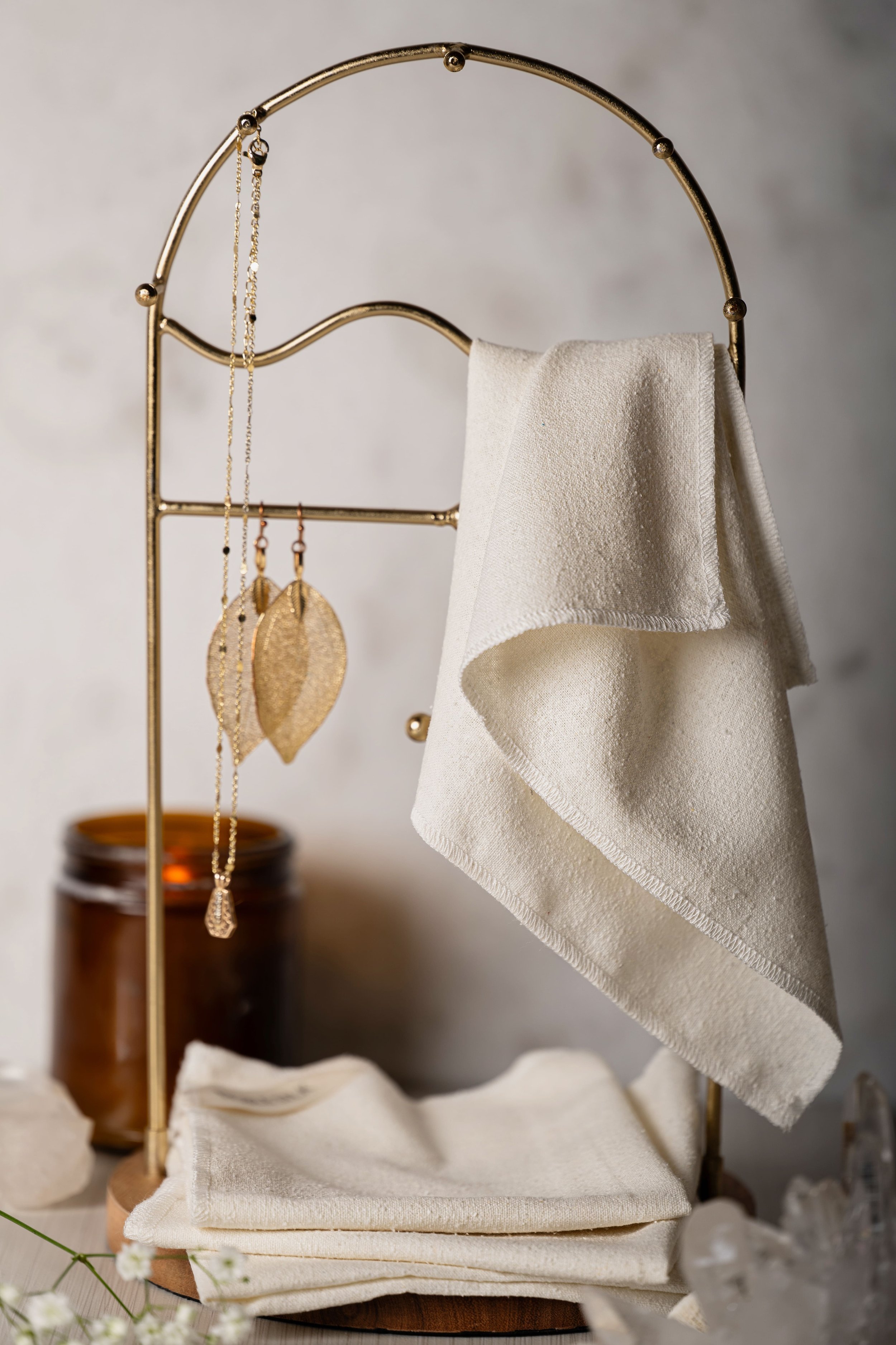 A gold jewelry holder with a white cloth and earrings hanging, set against a neutral wall with a brown jar in the background and some white cloth at the base.