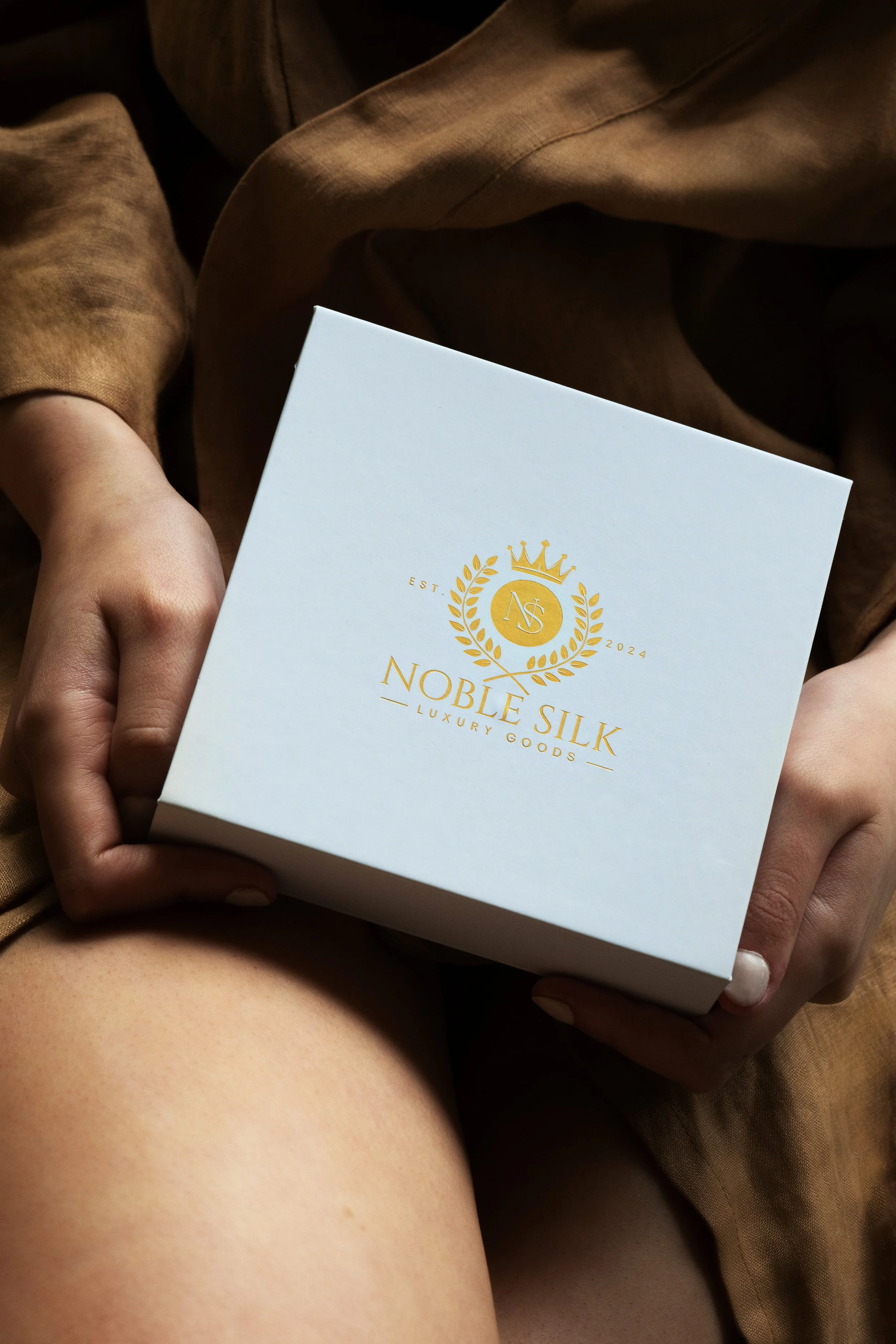 Person holding a white box with gold branding that reads 'Noble Silk Luxury Goods' and a crest logo.