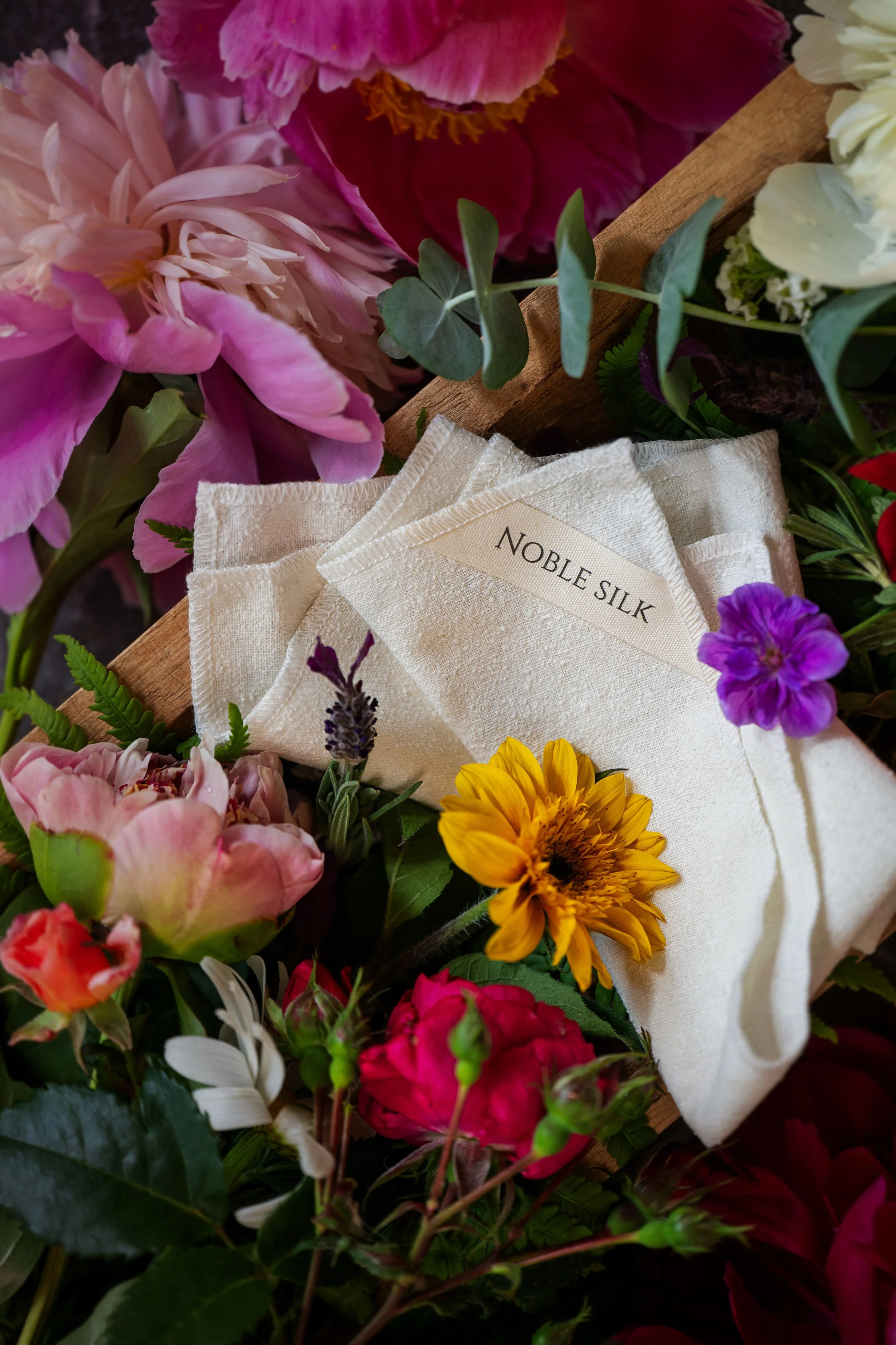 A collection of colorful flowers surrounding a white cloth with the label 'Noble Silk' on it.