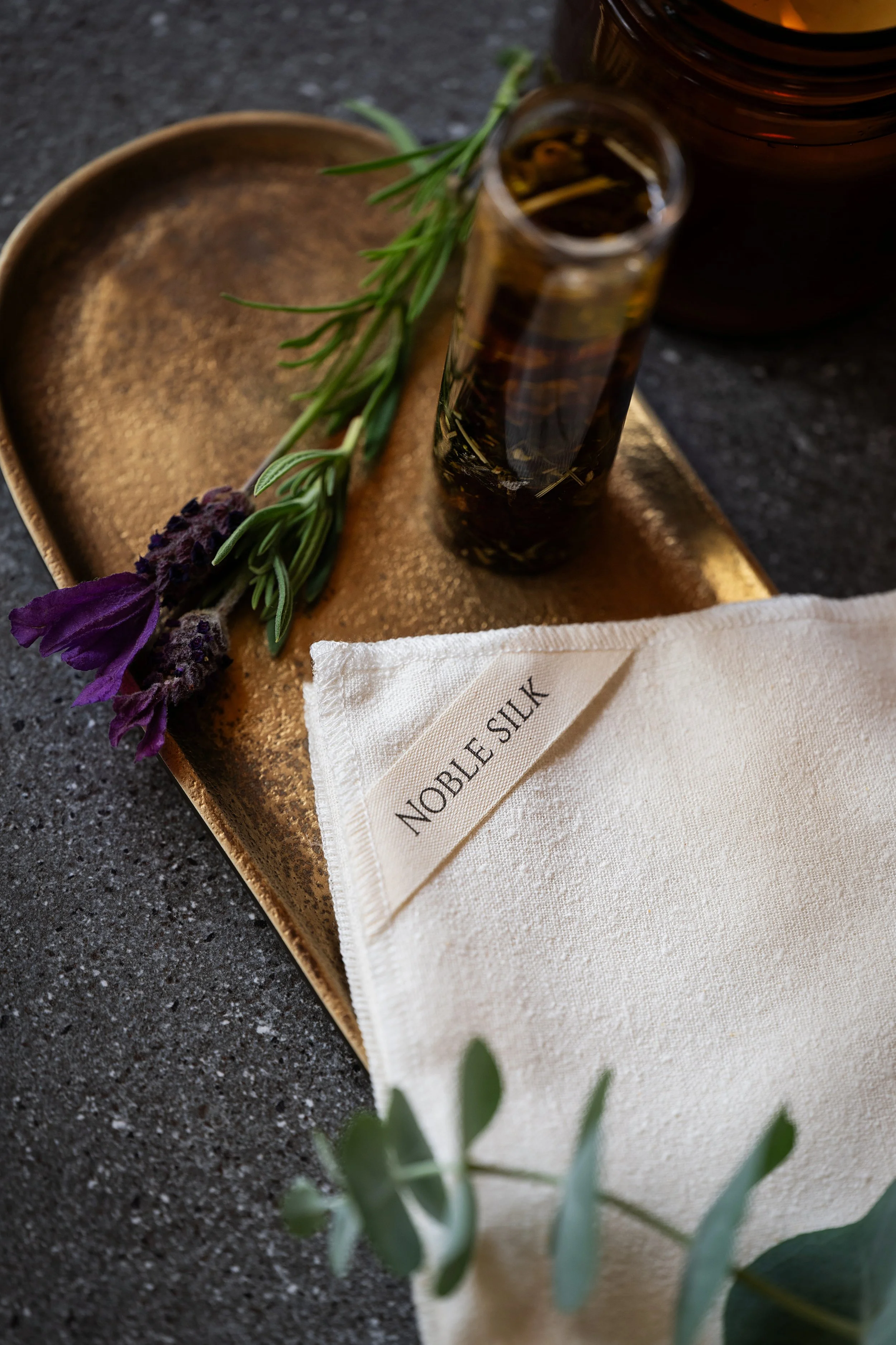 A rustic gold tray with a sprig of lavender and rosemary, a small glass bottle of essential oil, a cloth napkin labeled 'Noble Silk,' and a dark amber bottle in the background.