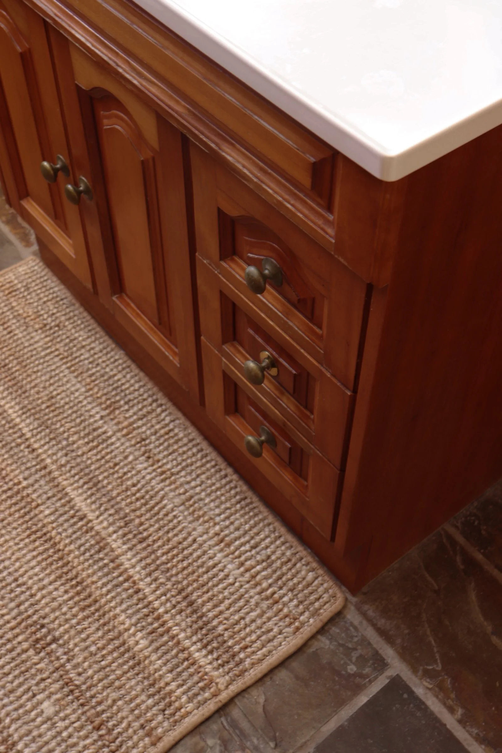Wooden bathroom vanity with multiple drawers and gold knobs, topped with a white countertop, next to a beige woven rug and tiled floor.