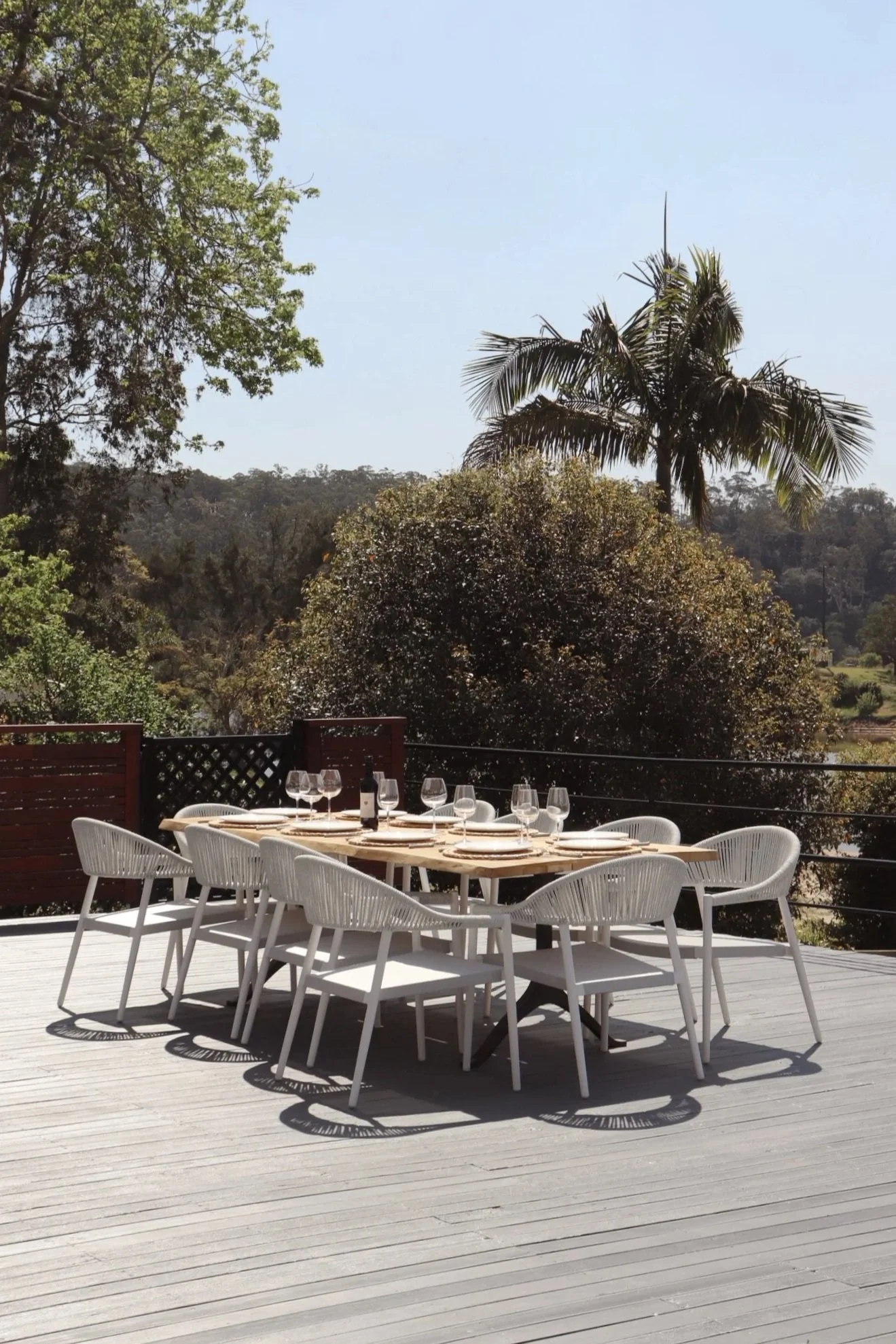 Outdoor dining table set with wine glasses, wine bottle, and plates on a deck with trees and a clear sky in the background.