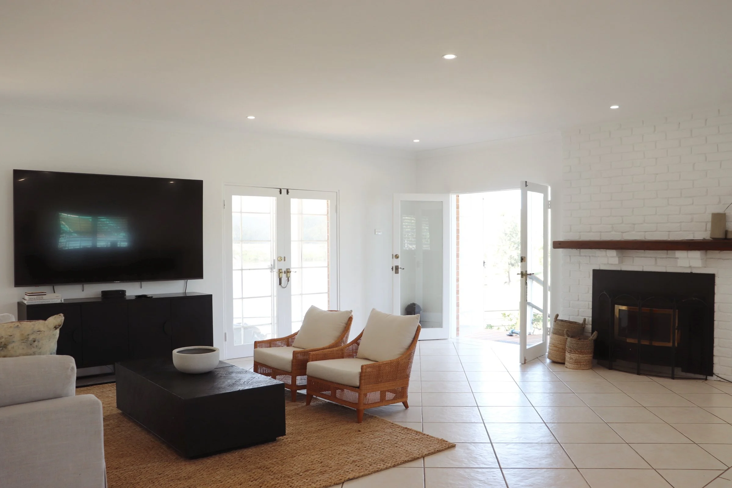 Bright living room with white walls, tiled floor, a large flat-screen TV on a black stand, wicker chairs with cushions, a coffee table, and a fireplace with a wooden mantel. Double doors and a glass door lead outside.